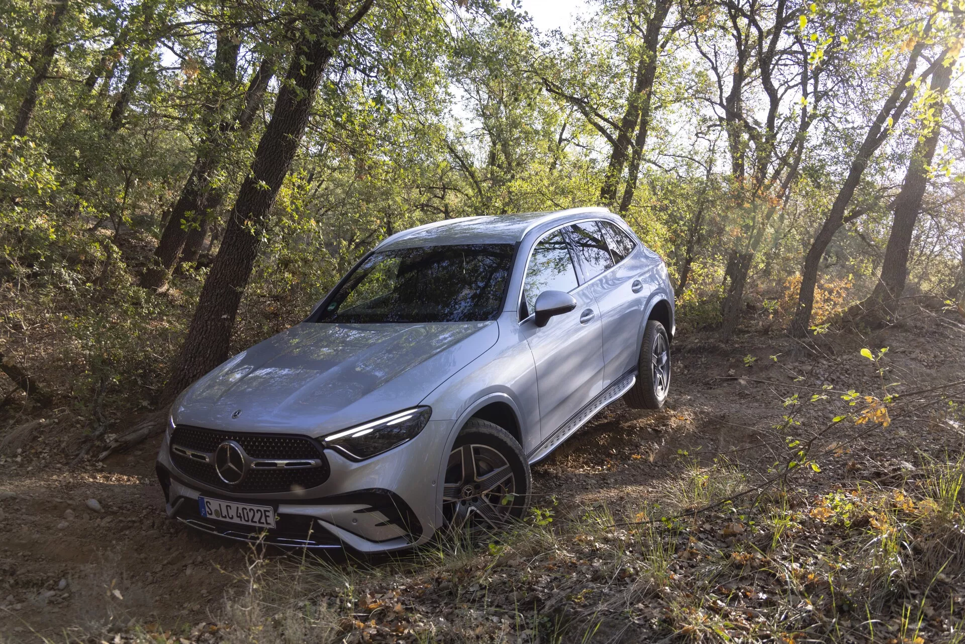 Slika srebrnega 2022 Mercedes GLC SUV, ki se nahaja zunaj v gozdnatem območju in prikazuje njegove terenske zmogljivosti. Vozilo je nagnjeno v levo, kar omogoča pogled na tri četrtine sprednjega in bočnega profila, vključno s sprednjo masko, ostrimi LED žarometi, konturiranim sprednjim odbijačem, pokrovom motorja, aluminijastimi kolesi s pnevmatikami za vse terene in gladkimi linijami. Naravna sončna svetloba se prestre skozi drevesa in poudarja moderen, aerodinamičen dizajn ter nakazuje prisotnost naprednih funkcij za pomoč pri vožnji, ki so nameščene za emblemom rešetke.