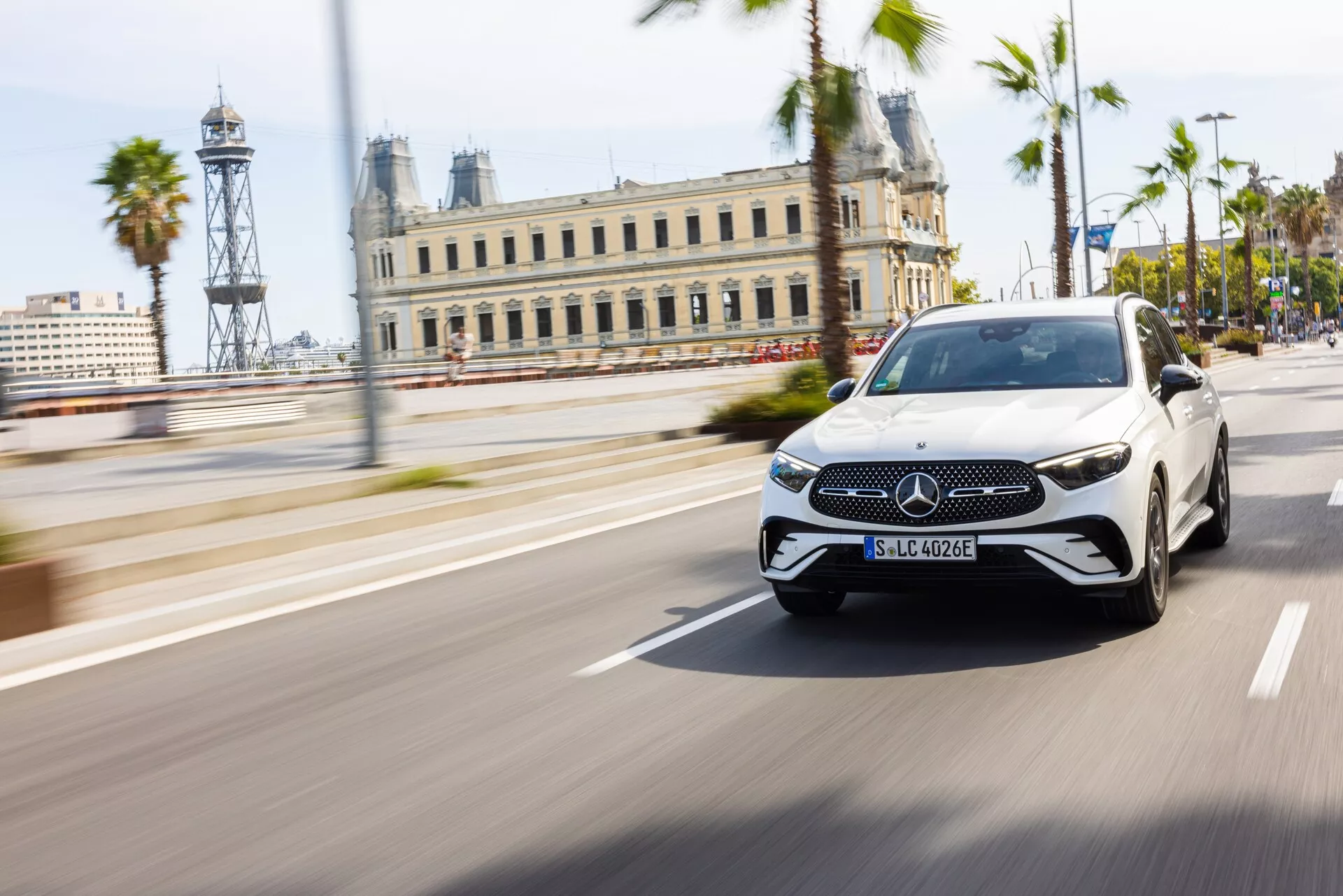 Mercedes-Benz GLC 2022 (254) luksuzna električna limuzina v beli barvi s črno streho, ujeta v gibanju v dnevnem urbanem okolju s palmami in zgodovinskimi zgradbami v ozadju, uporabljajoč tehniko panoramiranja za ohranjanje vozila ostrega proti zamegljenemu ozadju. S prepoznavnim emblemom Mercedes-Benz na rešetki, senzorji za pomoč vozniku, tankimi dnevnimi lučmi, elegantnimi LED žarometi in vgrajenimi smerniki v ogledalih.