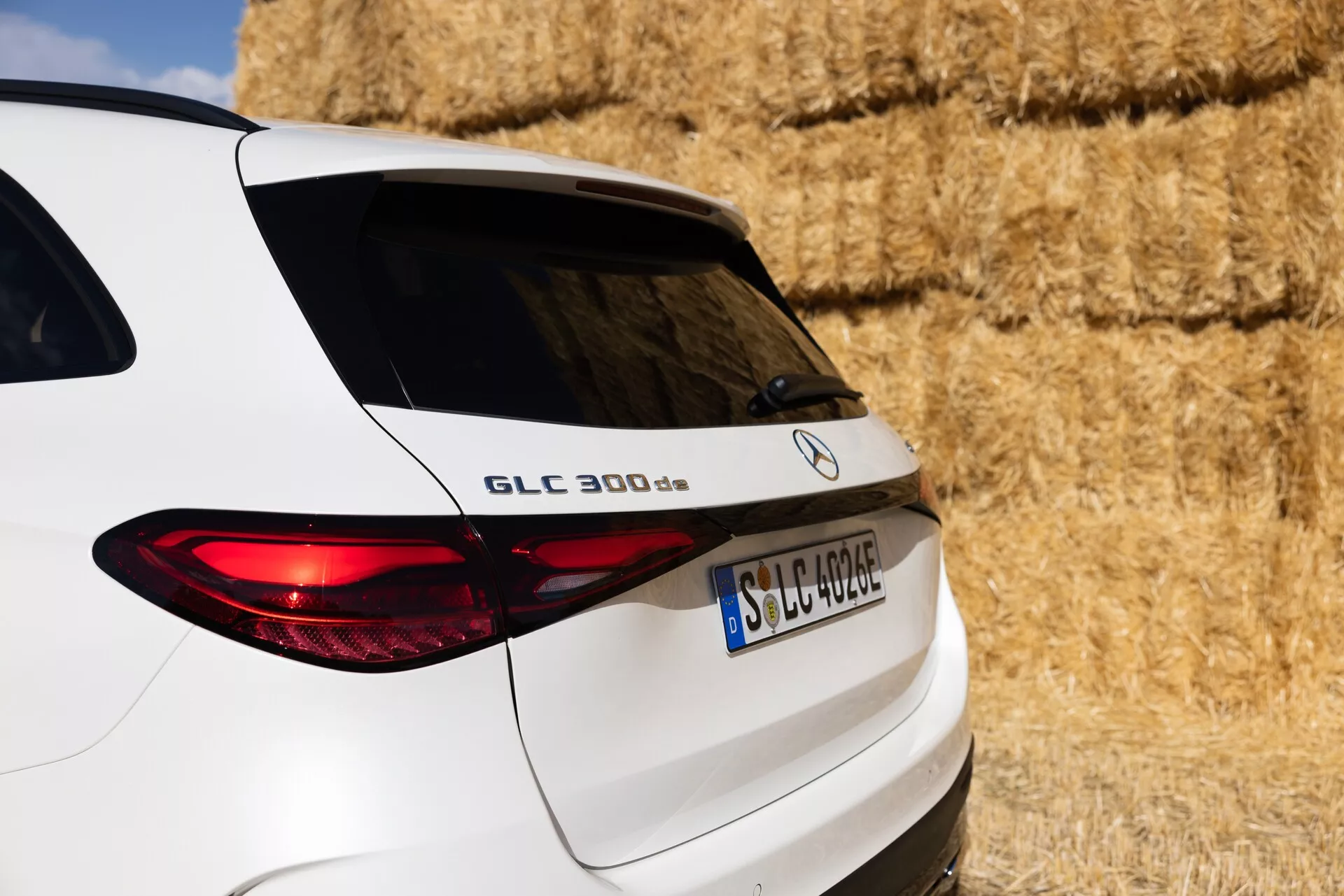 Od blizu, tričetrtinski zadnji pogled na bel Mercedes-Benz GLC 300 d SUV, ki prikazuje desno stran zadka, vključno s prtljažnimi vrati, osvetljenim zadnjim lučem in delom zadnjih oken. Vozilo je parkirano v ruralnem okolju z balami sena v ozadju. Vidne lastnosti vključujejo ime modela 'GLC 300 d', logotip Mercedes-Benz in registrsko tablico v evropskem formatu, ki poudarjajo elegantne oblikovne linije in moderno osvetlitev vozila.