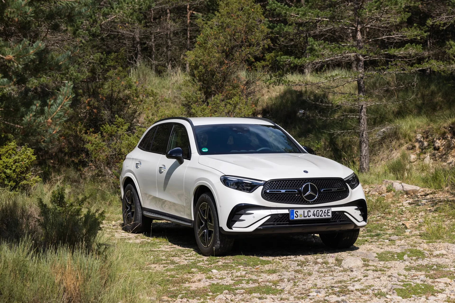 Slika SUV Mercedes-Benz GLC 2022 v naravnem okolju, postavljenega na makadamski ali peščeni poti z vegetacijo v ozadju. Vozilo je posneto iz tričetrtinskega sprednjega pogleda, ki prikazuje sprednjo masko z velikim emblemom, ostre LED žaromete in del bočne karoserije. SUV ima večkraka črna lita platišča, stranska ogledala z vgrajenimi smerokazi in dvigajočo se linijo pasu, ki vodi do športnega, ožanega zadnjega dela. Karoserija je v trdnem beli barvi, slika pa nakazuje, da je ta model del elektrificirane ponudbe Mercedes-Benz, kar nakazuje značko 'EQ' na sprednjem blatniku.