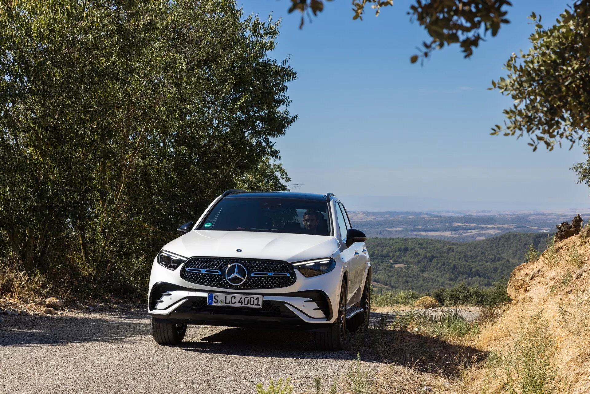 Moderen Mercedes-Benz GLC 2022 je nameščen na ozki cesti z naravno, hribovito kuliso, ki ponuja pogled na dolino ali ravnino. Prednji znak in oblikovanje rešetke avtomobila sta jasno vidna, posneta iz nizke čelne perspektive, rahlo levo, kar poudarja prednjo rešetko, žaromete in del pokrova motorja. LED žarometi, značilna rešetka z velikim emblemom in športna zasnova odbijača poudarjajo napredne in elegantne funkcije avtomobila. Dnevni posnetek z naravno svetlobo vzbuja občutek miru, saj prikazuje napredne sisteme za pomoč vozniku, ki jih nakazuje emblem. Eleganten aerodinamični profil je poudarjen z linijami pokrova motorja, ki se združujejo z izklesanimi boki, posneti na razdalji približno 3-5 metrov od kamere.