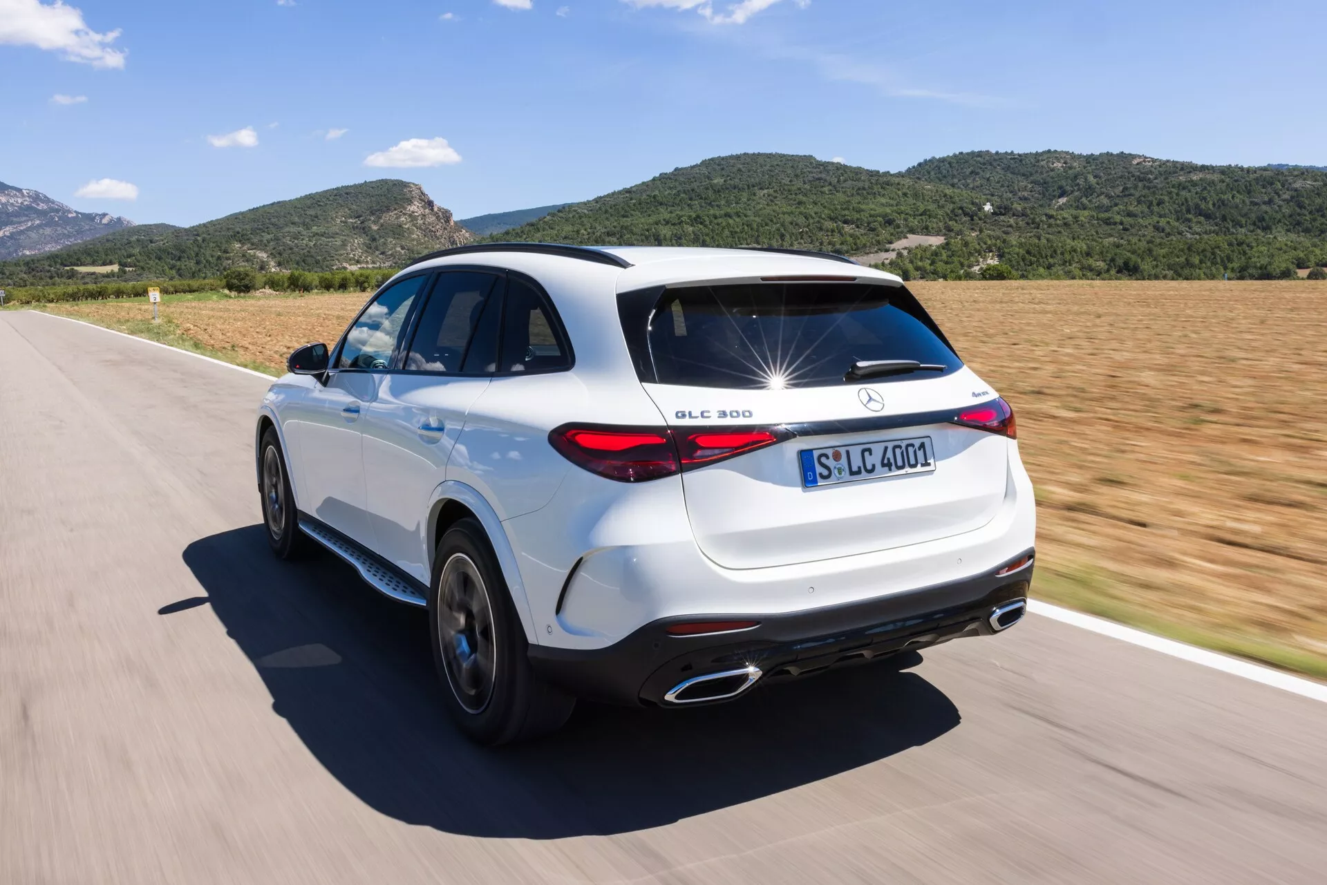 Visok zadnji tričetrtinski kot vidika belega Mercedes-Benz GLC 300 SUV na cesti. Slika prikazuje značilnosti oblikovanja, kot so prepoznavni znaki, LED zadnje luči, kromirani izpušni zaključki, petkraka aluminijasta platišča in značilen strešni spojler. SUV je v gibanju ob sončnem dnevu, kar poudarja njegove dinamične zmogljivosti in športno estetiko.