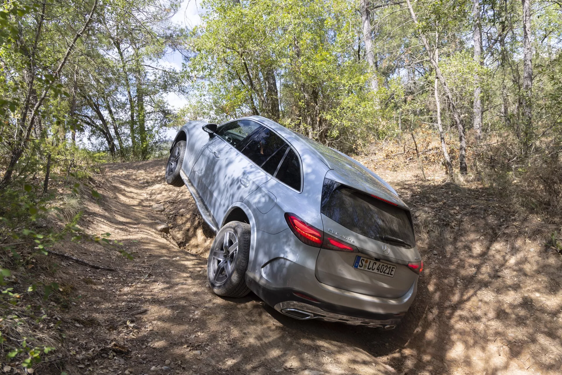 Visok, trikraten pogled zadnjega dela SUV-ja Mercedes GLC 2022 na makadamski poti v gozdnatem območju, ki prikazuje njegovo zmogljivost pogona na vsa kolesa, eleganten dizajn s panoramsko streho, LED zadnjimi lučmi, kovinski lak, zatemnjena stranska okna z vgrajenimi smerniki, vgrajeni ročaji vrat in velika platišča iz lahke litine.