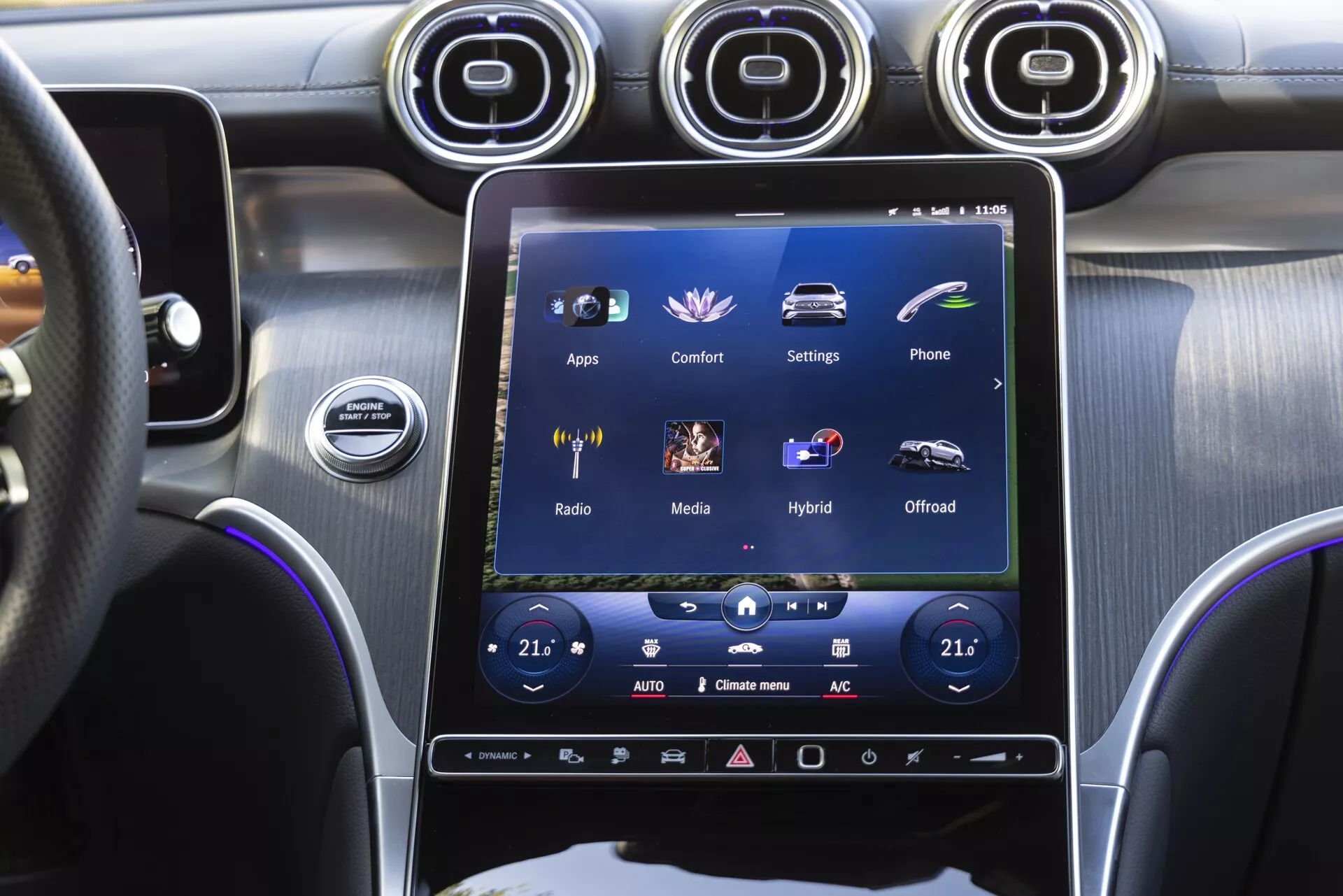 Centralna konzola Mercedes-Benz GLC 2022, gledana s sedeža voznika, osredotočena na velik navpično usmerjen zaslon na dotik infotainment sistema, ki prikazuje možnosti, kot so 'Aplikacije,' 'Udobje,' 'Nastavitve,' skupaj z dvopodročno klimatsko kontrolo, kovinskimi zračniki, gumbom za zagon/prost in premium mešanico ščetkanega kovinskega in visokosvetlečega črnega oblazinjenja.