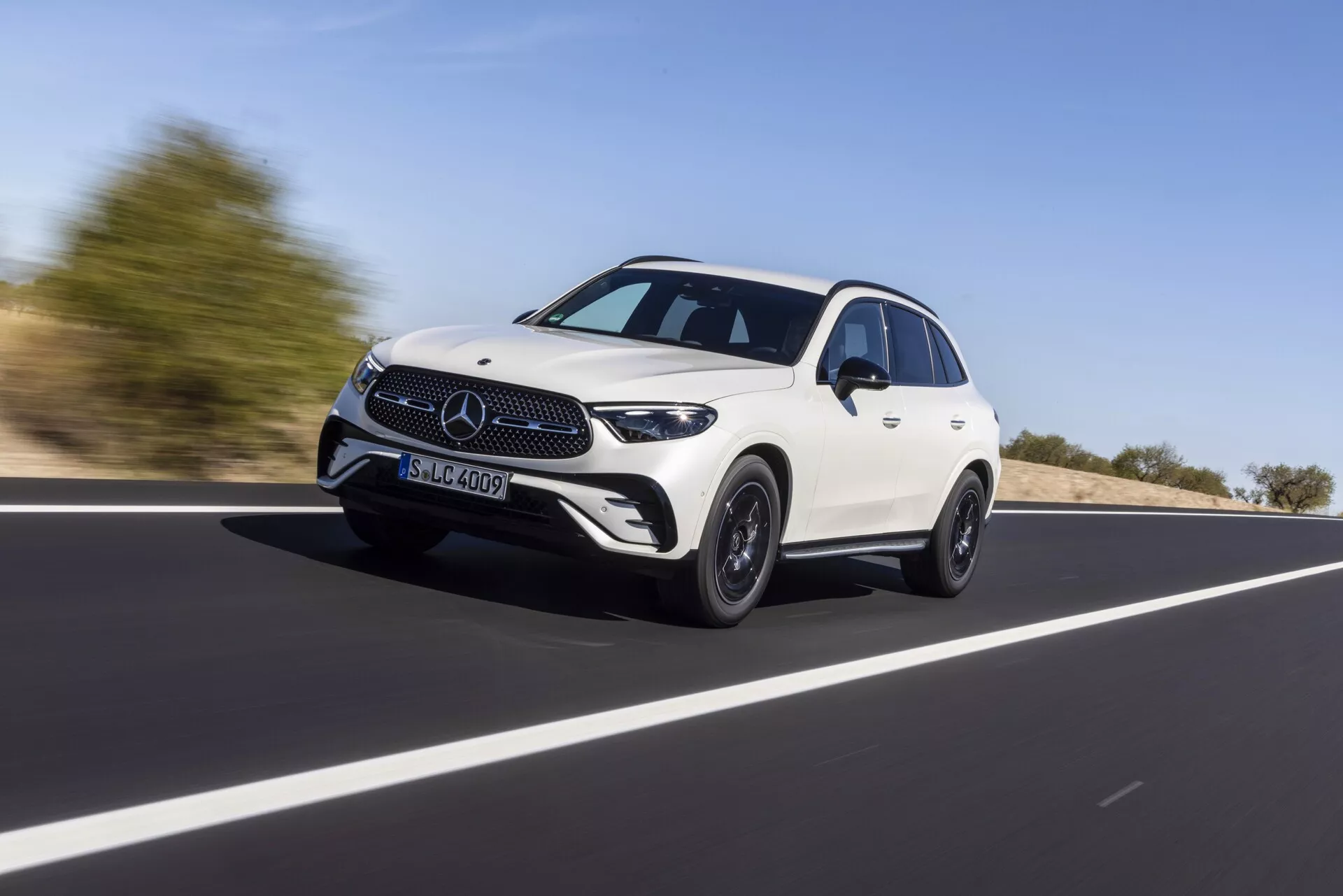 Mercedes GLC 2022 (254) bel štirivratni crossover SUV v gibanju na odprti cesti, zajet iz nizkega kota, ki poudarja njegovo višino in držo. Vozilo se giblje diagonalno z leve proti desni, s poudarkom na sprednji maski, žarometih, LED dnevnih lučeh in spodnjem prednjem odbijaču. Bočni profil razkriva izklesano strešno linijo, oblikovne linije, razširjene koloteke in platišča iz zlitine, ki združujejo razkošje in športnost. Zamegljeno ozadje nakazuje hitrost, pod jasnim dnevnim nebom.