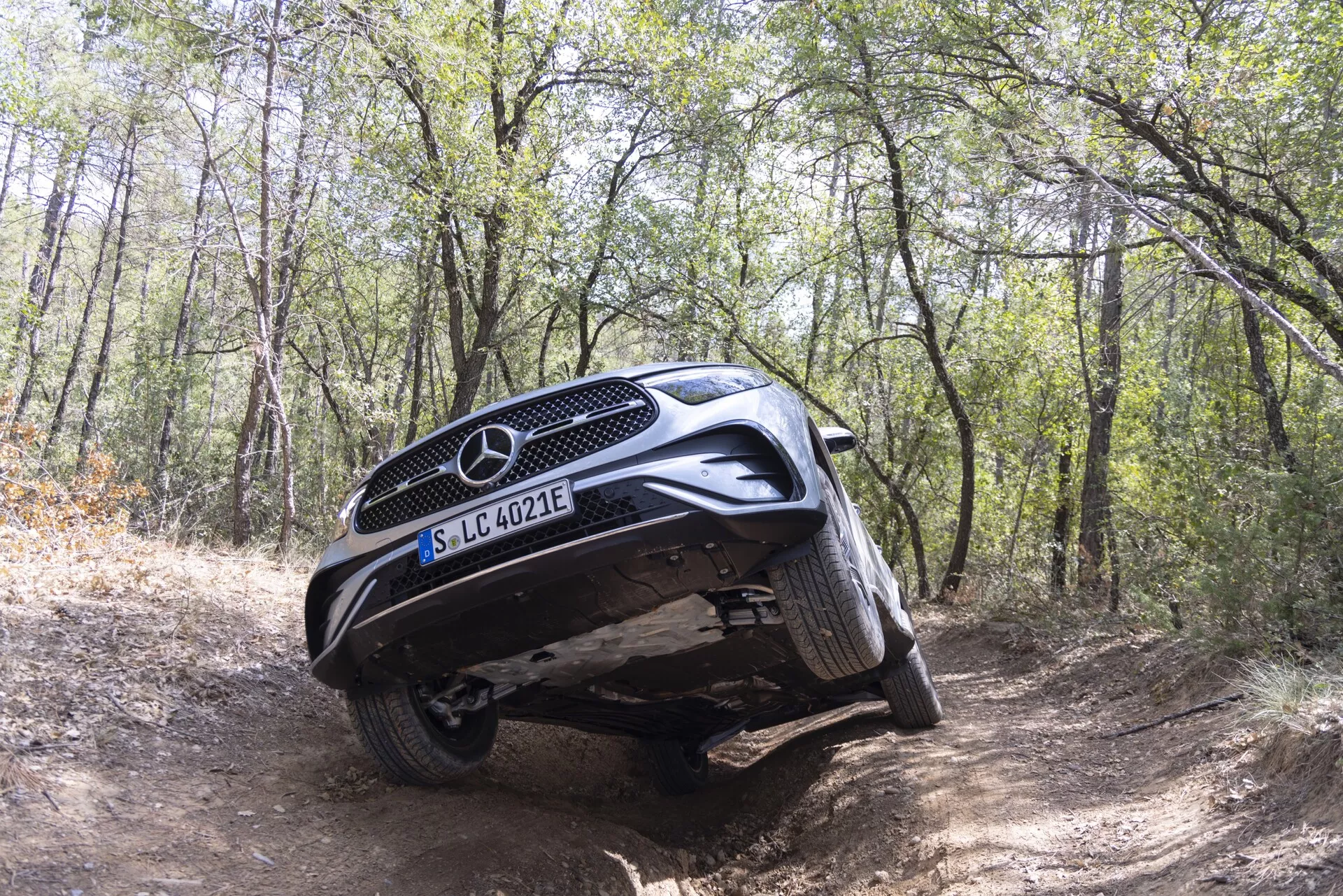 Mercedes-Benz GLC 254 iz leta 2022, zajet v terenskem okolju v gozdu, prikazuje njegove zmogljivosti na zahtevnem terenu. Nizkokotna, rahlo prednja fotografija poudarja sprednjo mrežo, zaščitno ploščo in artikulacijo vzmetenja z dvignjenimi levimi kolesi nad grbino in desnimi kolesi v jarku. To poudarja odmik od tal in terenske zaščitne lastnosti vozila. Znak EQ nakazuje električno ali hibridno tehnologijo. Naravna dnevna svetloba, brez umetne osvetlitve.