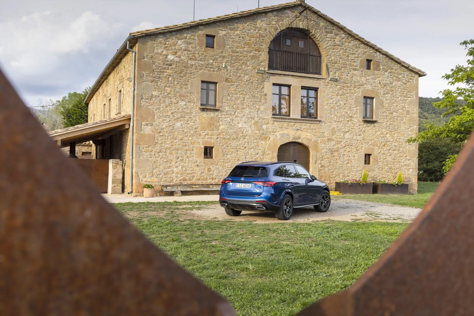 Mercedes GLC 2022 (254) parkiran na travnati površini s tradicionalno kamnito hišo v ozadju, prikazuje zadnji in delni stranski profil z LED zadnjimi lučmi, strešnim spojlerjem, strešnimi nosilci, zadnjimi parkirnimi senzorji, litoželeznimi platišči in zarjavelimi železnimi elementi v ospredju.