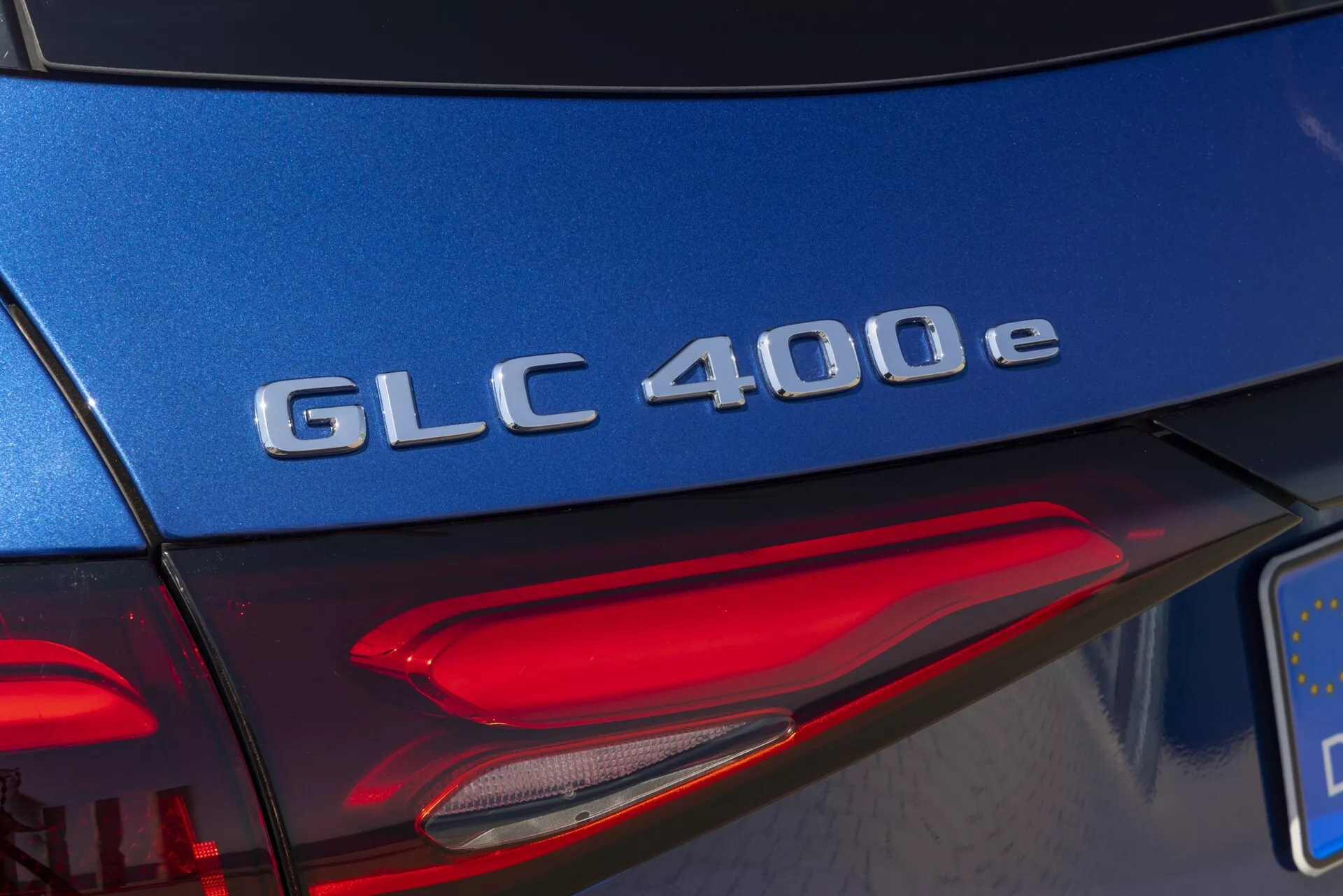 Podrobnost zadnjega dela Mercedes-Benz GLC 400e 2022, s bližinskim posnetkom oznake modela v kromu na bogato modri prtljažni vratih, z diagonalo usmerjenim kotom, ki prikazuje del leve zadnje luči z LED elementi pri dnevni svetlobi.