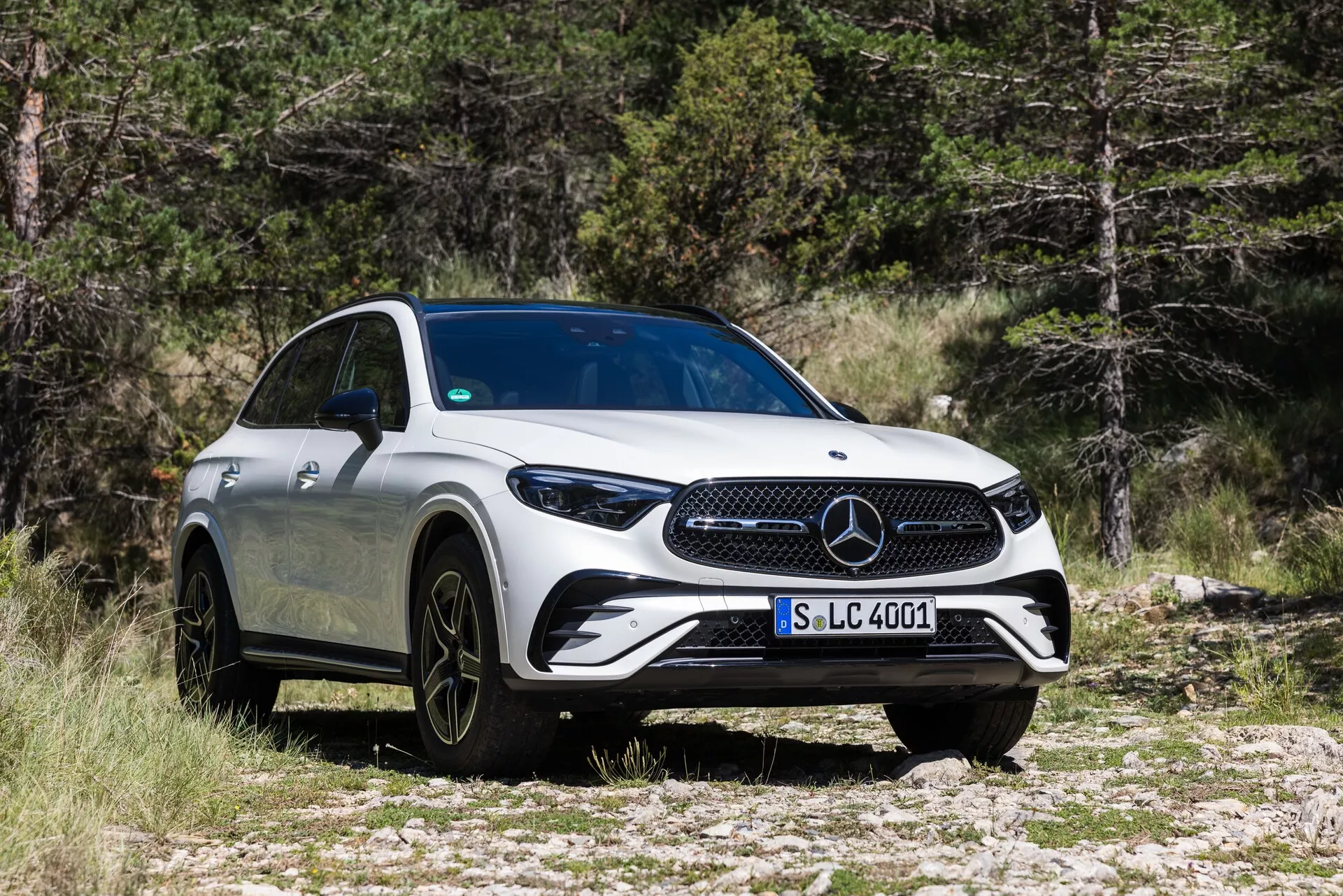 Trikratni sprednji pogled na Mercedes-Benz GLC 2022, ki poudarja njegovo izrazito masko, elegantne LED žaromete, velika črna platišča iz več krakov in aerodinamično belo karoserijo. Vozilo je parkirano na skalnatem terenu z drevesi v ozadju, kar poudarja njegove SUV zmogljivosti in luksuzni videz.