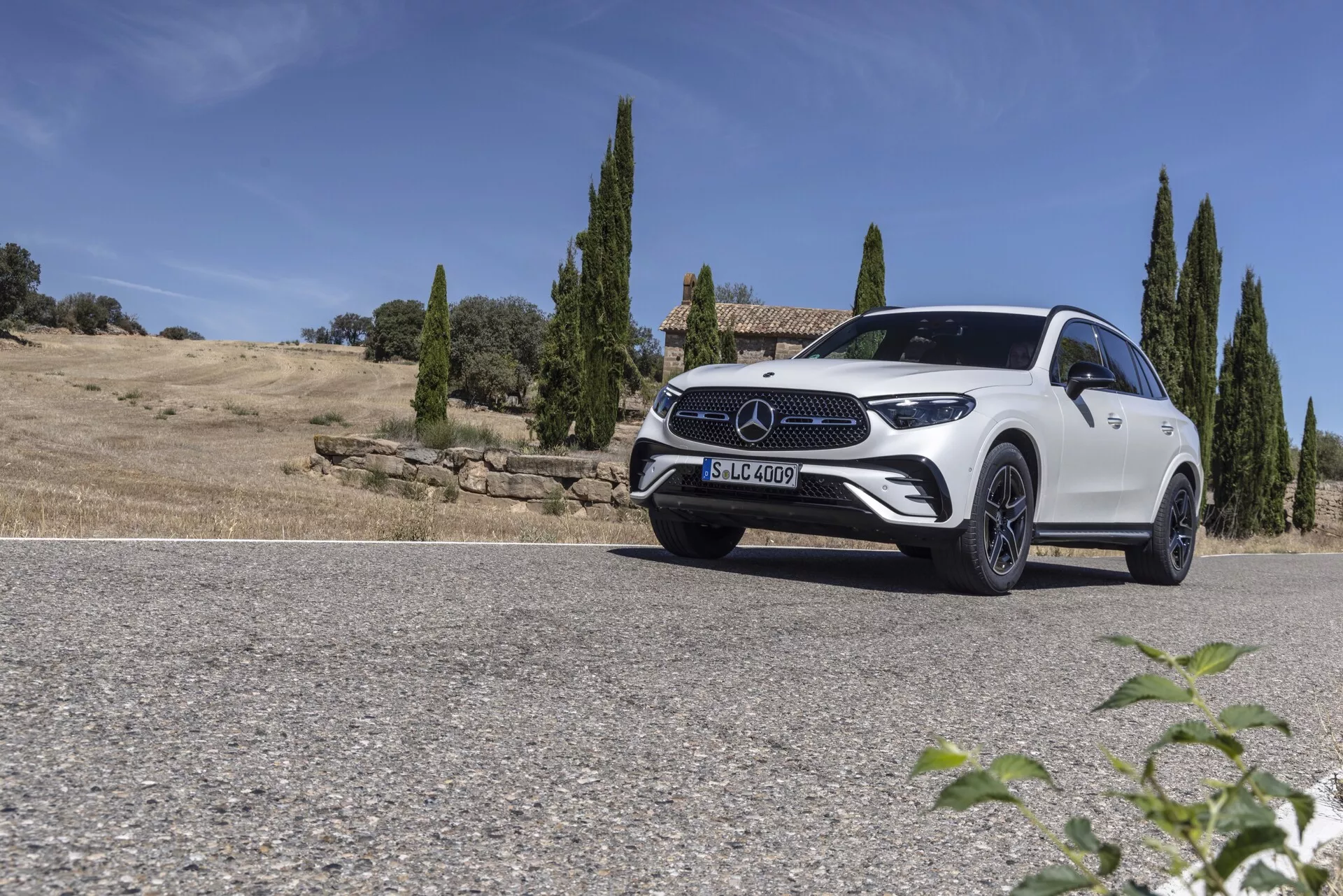 Nizek kotni posnetek belega SUV-ja Mercedes GLC 2022, parkiranega na cesti proti naravni pokrajini s čistim nebom in cipresami v ozadju. Sprednja četrtina vozila je ujeta s strani voznika, kar prikazuje elegantne LED žaromete, izrazito sprednjo mrežico z velikim emblemom, športna lita platišča in konturirano pokrov motorja. Rahlo nagnjena strešna črta in dobro definirana ramenska črta poudarjata dizajn, ki združuje aerodinamiko in robustnost. Svetla sončna svetloba ustvarja sence in svetlobne učinke, kar poudarja luksuzno in moderno estetiko vozila.