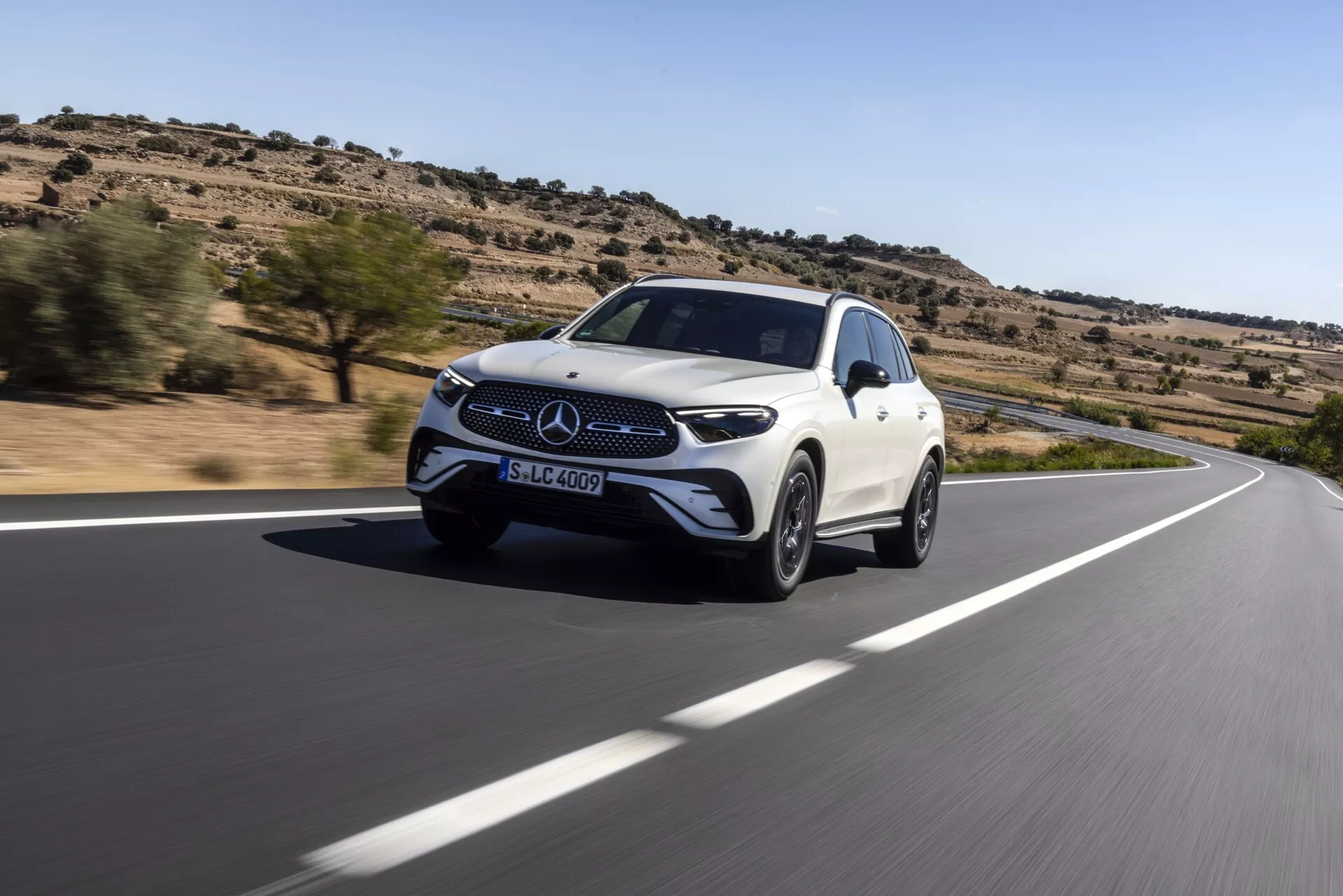 Dinamičen posnetek iz nizkega kota belega Mercedes-Benz GLC SUV iz leta 2022 med vožnjo po ovinkasti cesti. Pogled s tri četrtine spredaj zajema sprednjo masko z emblemi Mercedes-Benz, žaromete in sprednje blatnike. Skladna aerodinamična zasnova in lita platišča poudarjajo hitrost in luksuz vozila. Vidno v puščavskem okolju z redko vegetacijo, vgrajeni sprednji senzorji in stranska ogledala avtomobila nakazujejo napredne sisteme za pomoč voznikom in varnostne funkcije.