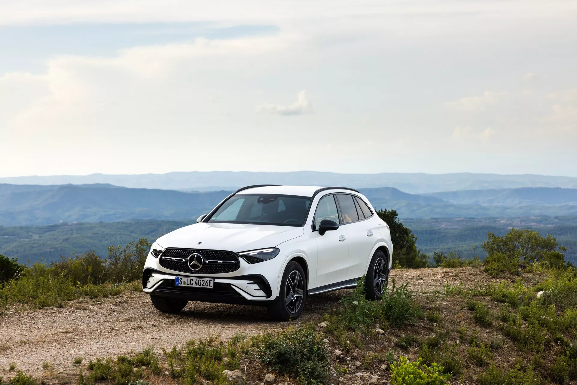 Stranski profil belega Mercedes GLC 2022 na neravni poti, poudarja držo, oddaljenost od tal in dizajnerske linije SUV-ja. Vidne lastnosti vključujejo sprednjo mrežo in značko, LED žaromete, lita platišča, stranska ogledala z vgrajenimi smerniki, aerodinamične linije karoserije, sijajni lak in vzorec pnevmatik. Naravna dnevna svetloba izboljša estetske in funkcionalne lastnosti vozila.