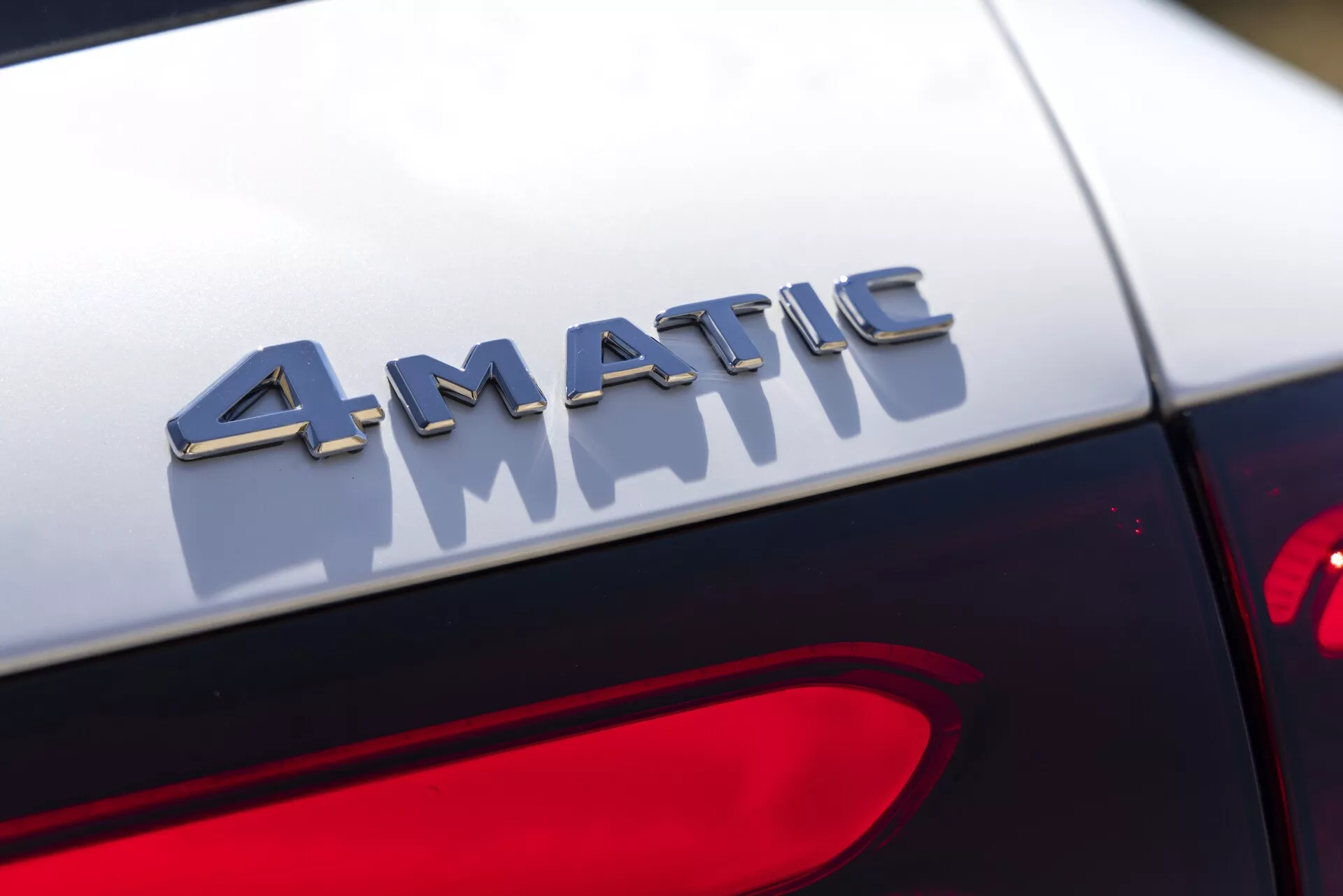Pogled od blizu na podrobnosti o znački 4MATIC na zadnjem delu avtomobila Mercedes GLC 2022. Fotografija zajame značko v ostri ostrini s kovinskimi črkami, ki odbijajo svetlobo, nameščeno tik nad zadnjo lučjo. Ozadje je zamegljeno, poudarja tridimenzionalni učinek značke in kontrast s temnim lakom avtomobila. Oblikovanje zadnje luči je delno vidno v spodnjem desnem kotu.