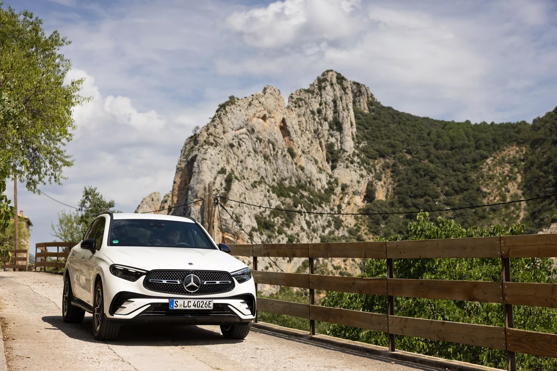 Beli Mercedes-Benz GLC 2022 parkiran na slikoviti cesti s pogledom z desne spredaj, ki prikazuje rešetko, žaromete in del desne strani vključno s sprednjimi sovoznikovimi vrati in kolesom. Registrska tablica 'SLC 400 e' nakazuje možnost priključnega hibrida ali električnega vozila. Ujet čez dan, bela barva in bleščeča površina avta odsevata naravno svetlobo, poudarjajoč veliki emblem Mercedes-Benza na rešetki. Kamnita gora in lesena ograja v ozadju nakazujeta podeželsko ali gorsko cesto, kar nakazuje luksuzni oddih ali raziskovanje na prostem.