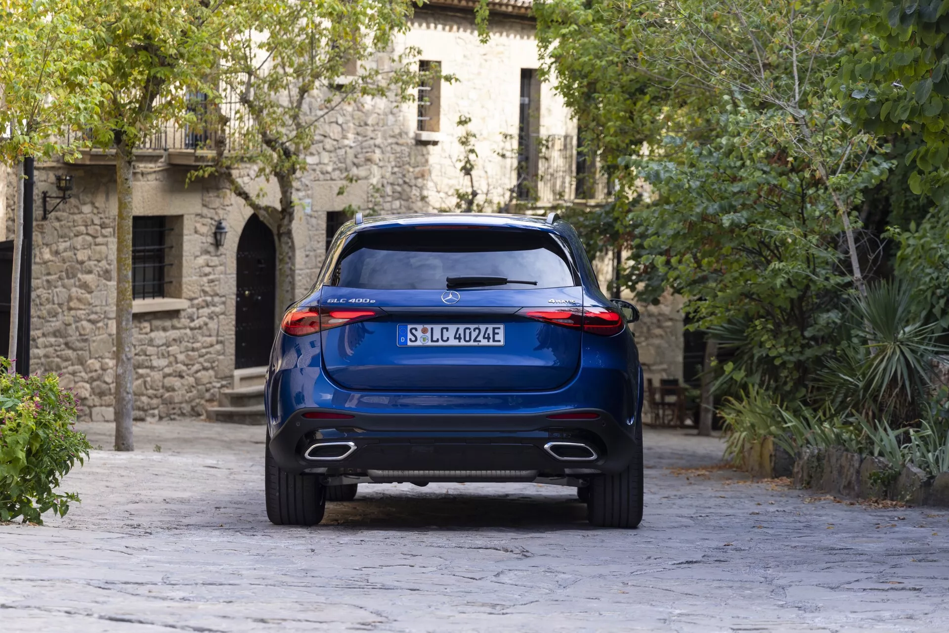 Zadnji kotni pogled na modri Mercedes GLC 2022 SUV iz nekoliko nizke perspektive, ki prikazuje visoko oddaljenost od tal, dvojne izpušne odprtine in elegantne zadnje svetlobne sklope. Vozilo, postavljeno sredi tega, stoji na ozadju evropske arhitekture in tlakovanega cestišča, kar poudarja njegovo modernost in možen električni pogonski sklop.