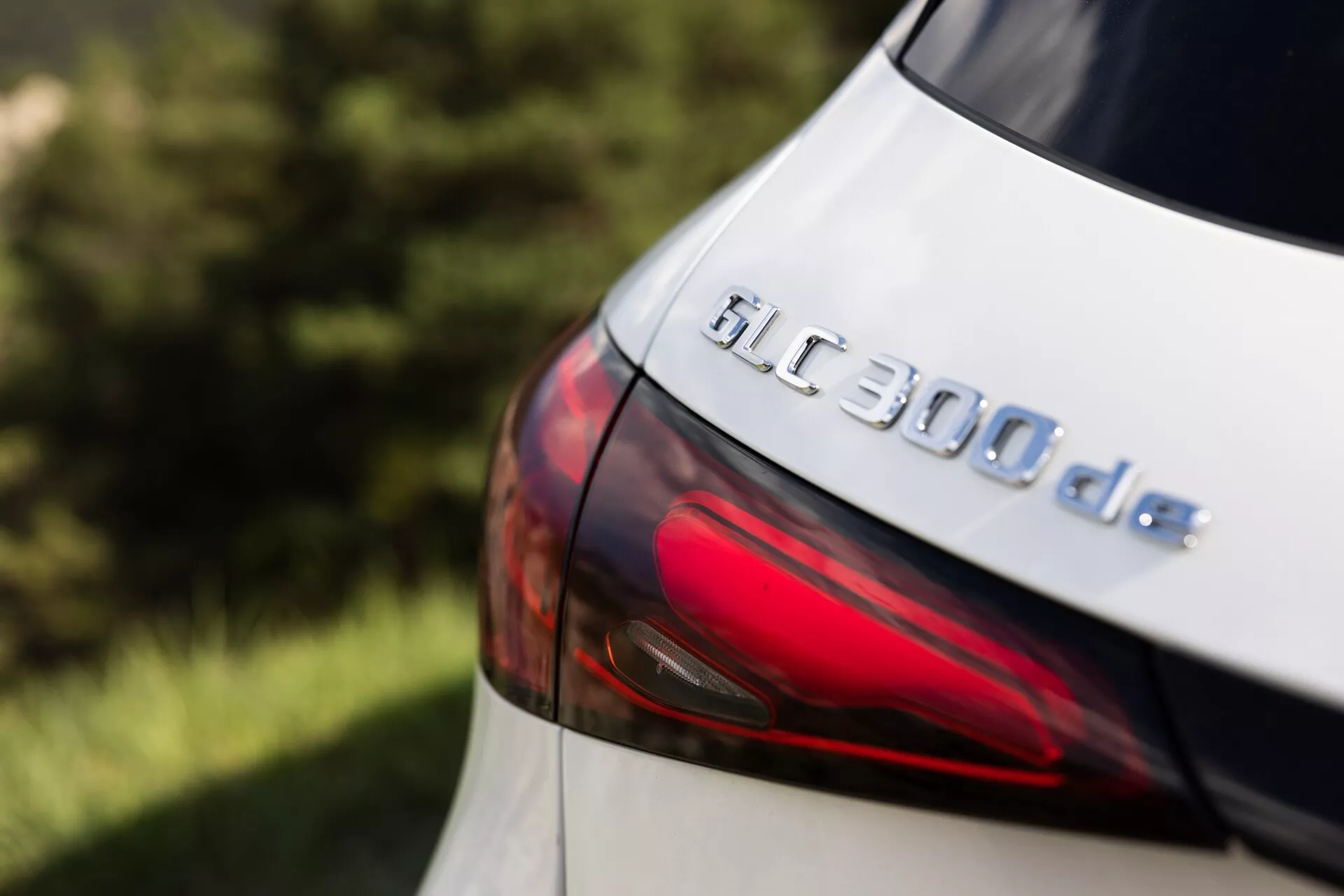 Podroben posnetek zadnjega dela Mercedes GLC 300de 2022, osredotočen na značko modela in del LED zadnje luči. Sliko, narejeno iz rahlo dvignjenega bočnega kota, poudarja značko GLC 300de in gladek obris sodobne oblike zadnje luči. Kovinska barva rahlo odbija svetlobo, kar nakazuje dobro vzdrževano površino, čeprav se natančna barva zdi svetla, verjetno bela ali srebrna. Plitva globinska ostrina ohranja značko v jasnem fokusu, hkrati pa zamegli ozadje in obrobna področja, poudarjajoč tehnološke in oblikovalske podrobnosti tega dizelsko-električnega hibridnega vozila.