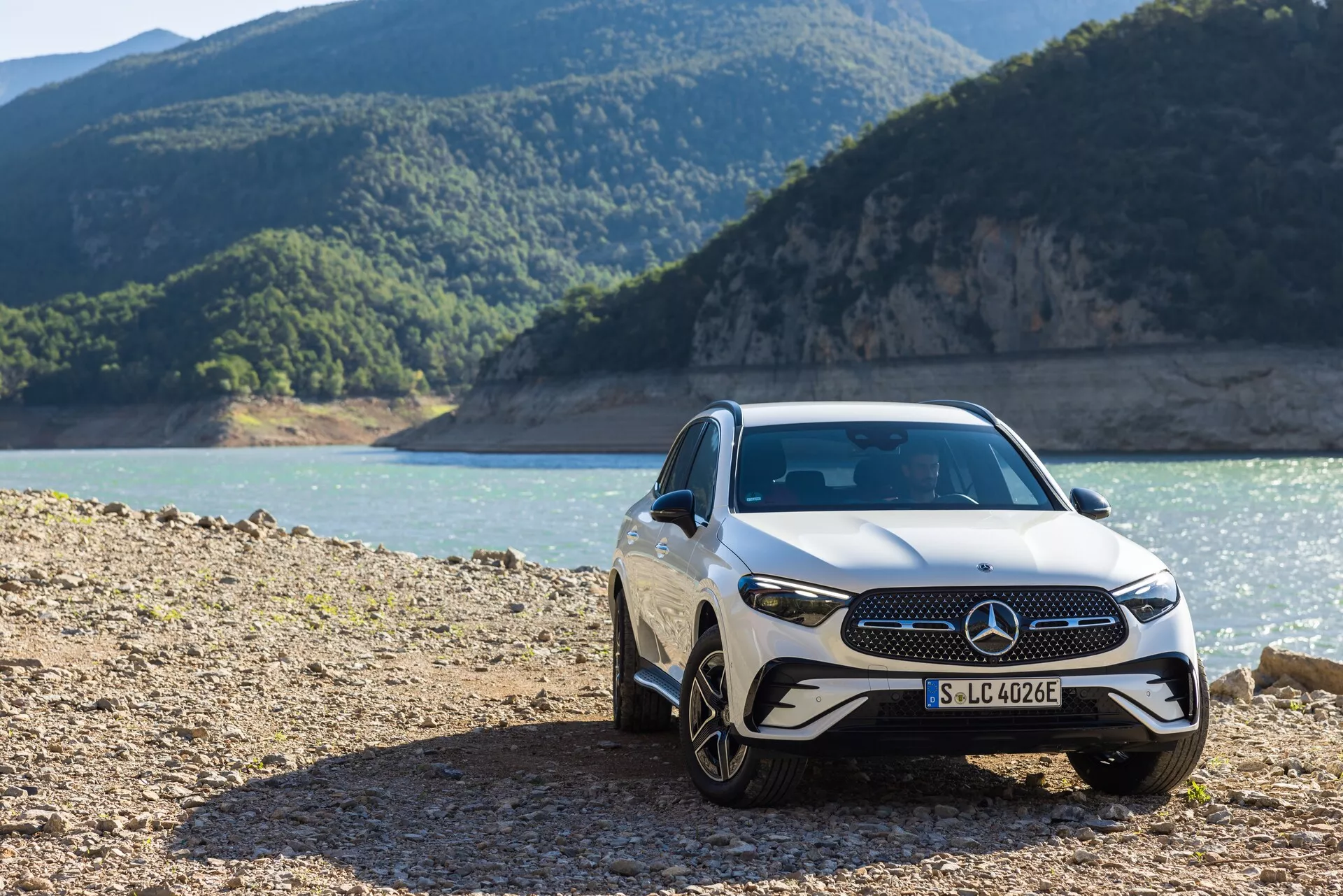 Prednji tridelni pogled na luksuzni avto Mercedes GLC 254 iz leta 2022, ki prikazuje sprednjo masko z vidnim emblemom, elegantne LED žaromete in aluminijasta platišča, parkiran v robustnem zunanjem okolju blizu obale z gorami v ozadju.