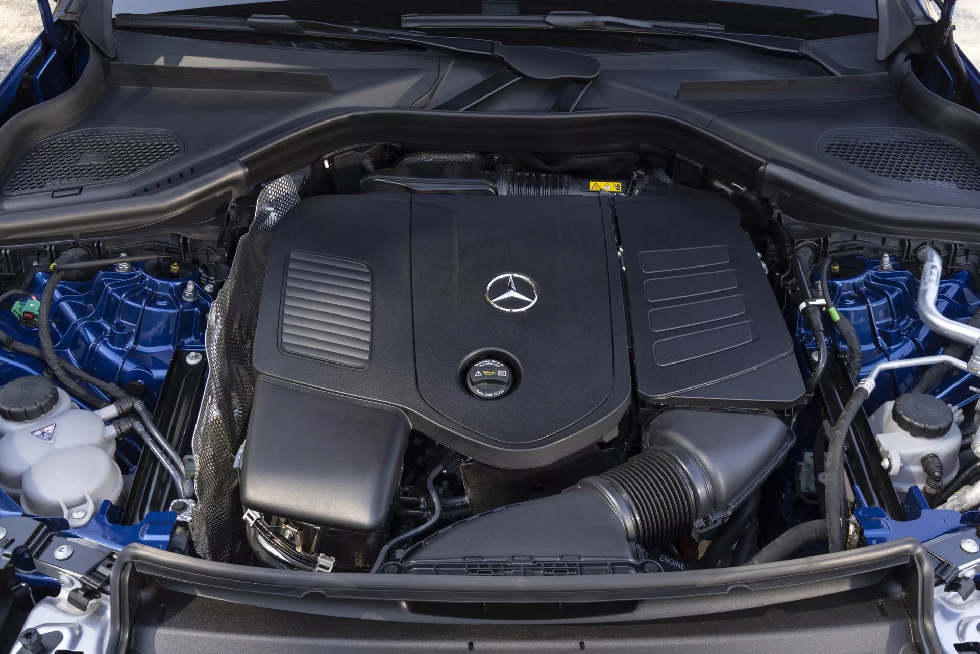 Podroben zgornji posnetek čistega in dobro vzdrževanega motorskega prostora Mercedes GLC 2022, ki prikazuje pokrov motorja z opaznim logotipom Mercedes-Benz, baterijo, rezervoarji za tekočine, sestavne dele hladilnega sistema in urejeno organizirane kabelske snope.