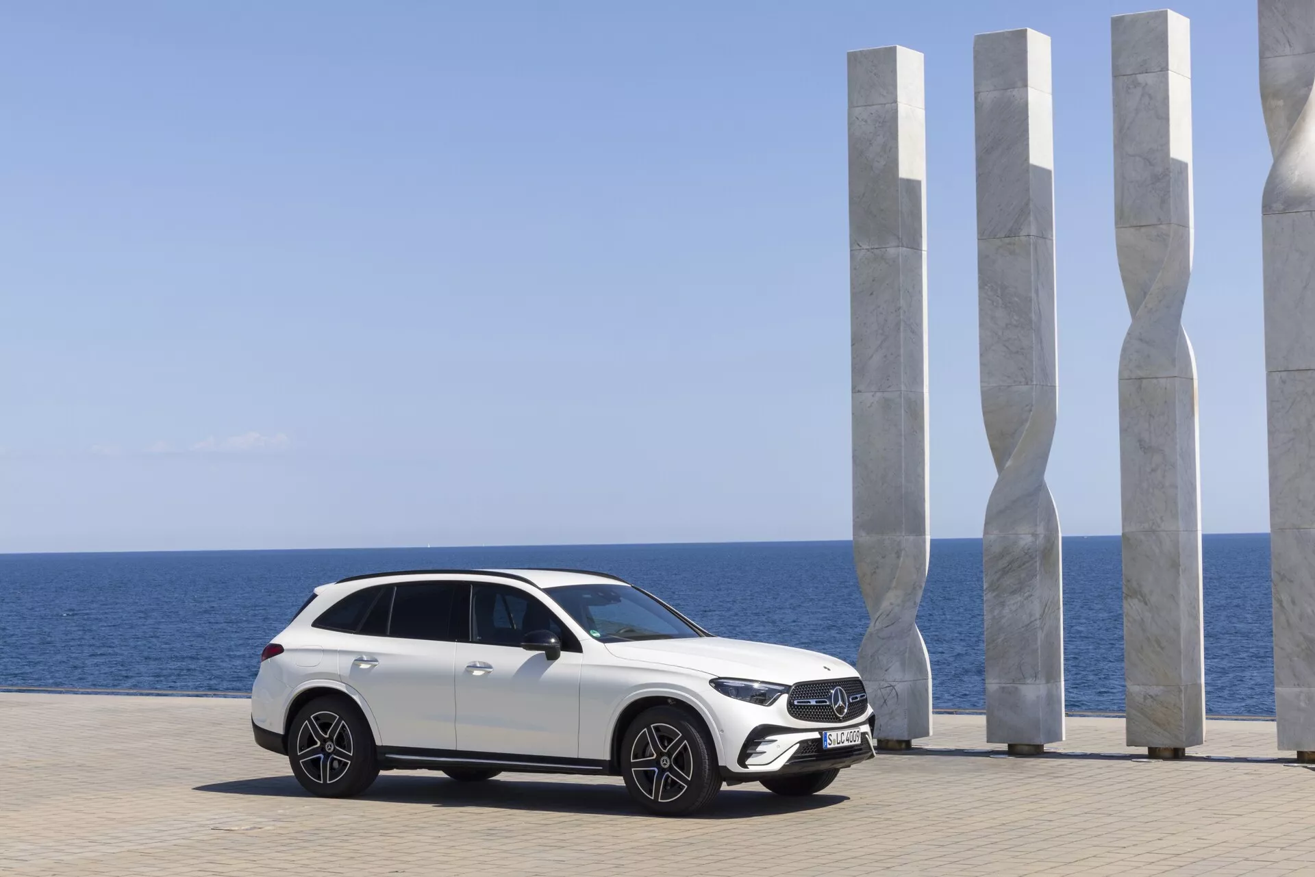 Moderni beli luksuzni Mercedes GLC 2022 parkiran s pogledom na slikovito obalo; stranski profil prikazuje eleganten dizajn z dvignjeno višino vožnje, velikimi aluminijastimi kolesi, naprednimi LED žarometi, aerodinamičnimi ogledali z vgrajenimi smerniki, poševno kupejsko streho, panoramsko streho in zatemnjenimi okni. Slikovito ozadje z modernimi belimi skulpturami, jasnim modrim nebom in mirnim morjem poudarja vrhunski avtomobilski privlak.