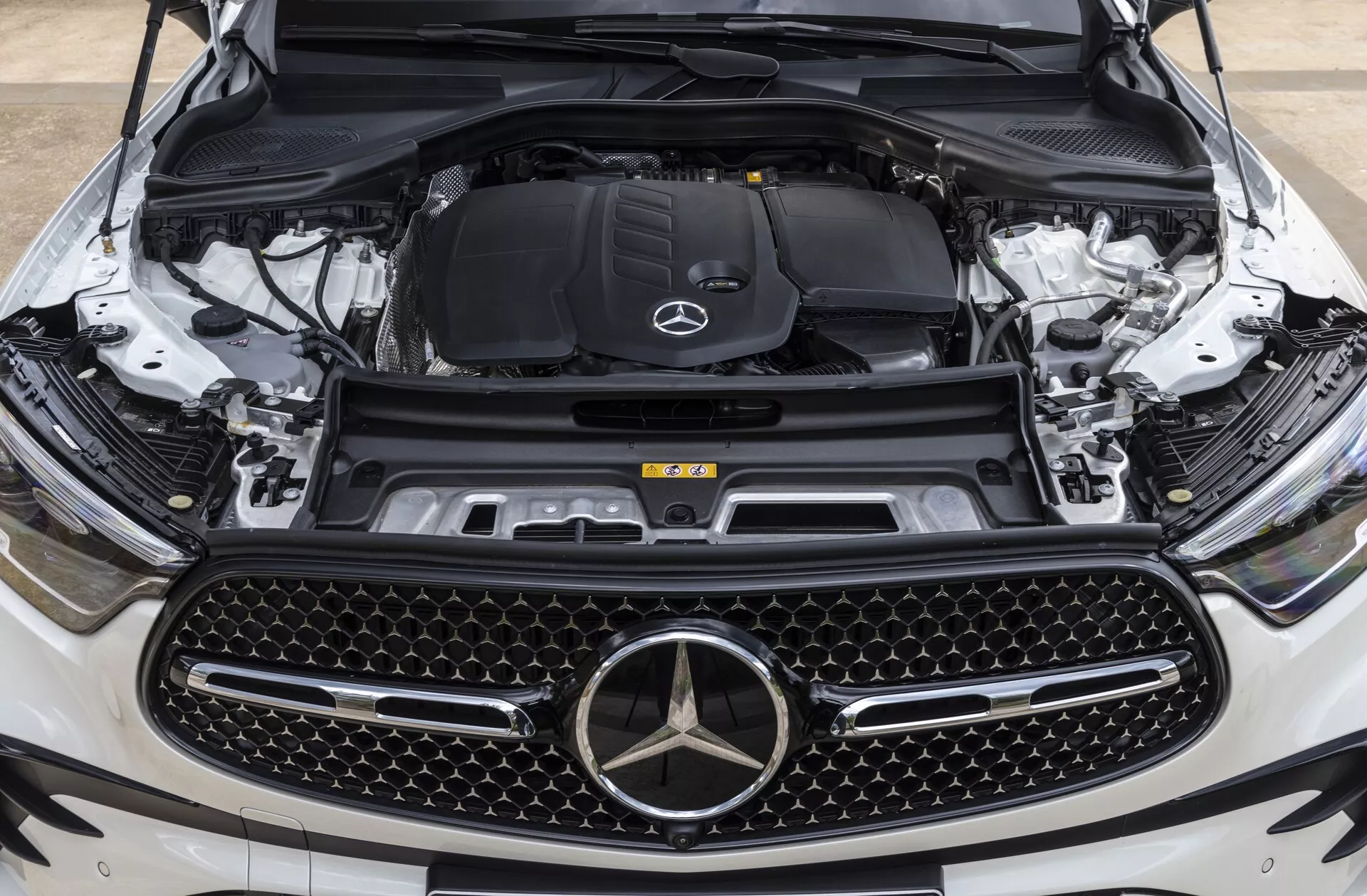 Povečan sprednji pogled na Mercedes-Benz GLC 2022 (254) z odprtim pokrovom motorja, prikazuje motorni prostor z vidika na višini oči, osredotočen na emblem blagovne znamke in mrežo, poudarja veliko mrežo z diamantnim vzorcem, emblem zvezde Mercedes-Benz, motorni prostor z logotipom in vidne komponente, kot so blok motorja, sistem za vnos zraka, rezervoar za hladilno tekočino, stolp vzmetenja in sprednja kamera, ki nakazujejo napredne tehnološke funkcije.