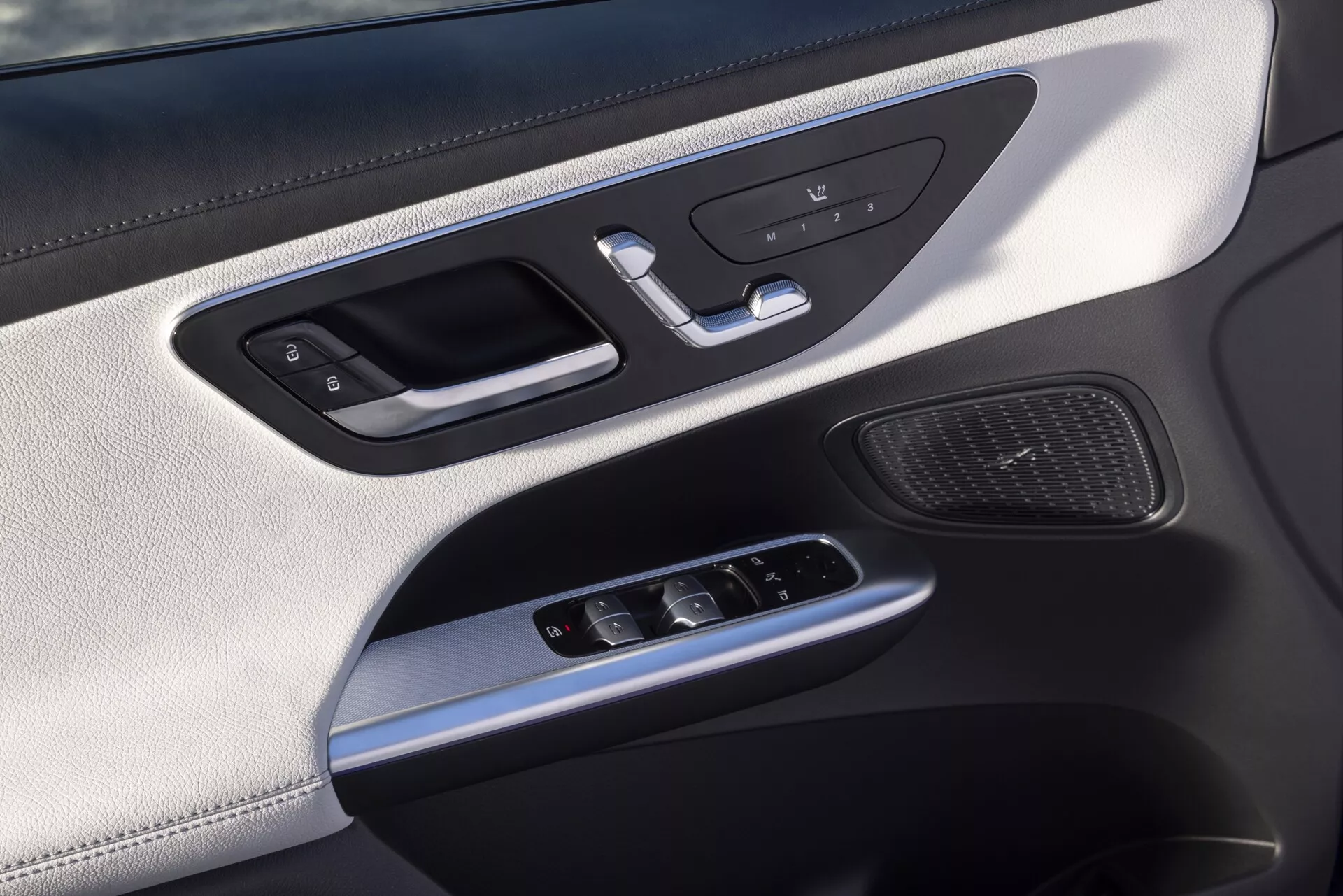 Pogled od blizu na notranjo oblogo vrat avtomobila Mercedes GLC 2022 254, osredotočen na stran voznika ali sopotnika, ki vključuje srebrno kljuko z integriranimi kontrolami za zaklepanje, gumbe za pomnilnik sedeža, stikala za električna okna, joystick za nastavitev stranskih ogledal, mrežo zvočnikov, razkošne materiale, vključno z usnjenim zgornjim delom, srebrnimi poudarki in ambientalno osvetlitvijo.