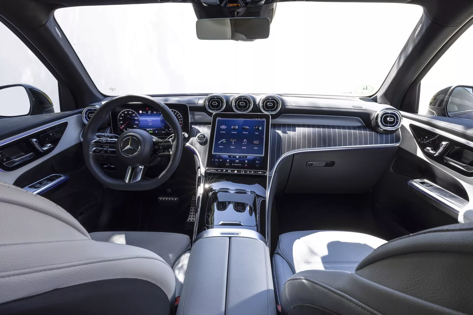 Notranjost Mercedes GLC 2022, gledano iz zadnjega sredinsko sedeža. Luksuzna kabina s dvobarvno shemo z kremnim usnjem in temnimi poudarki. Armaturna plošča ima eleganten leseni dekor z velikim vertikalnim zaslonom na dotik infotainment sistema, zračniki in kontrolnimi gumbi. Vidni sta tudi večfunkcijski usnjen volan s kovinskimi poudarki in digitalni instrumentni plošči.