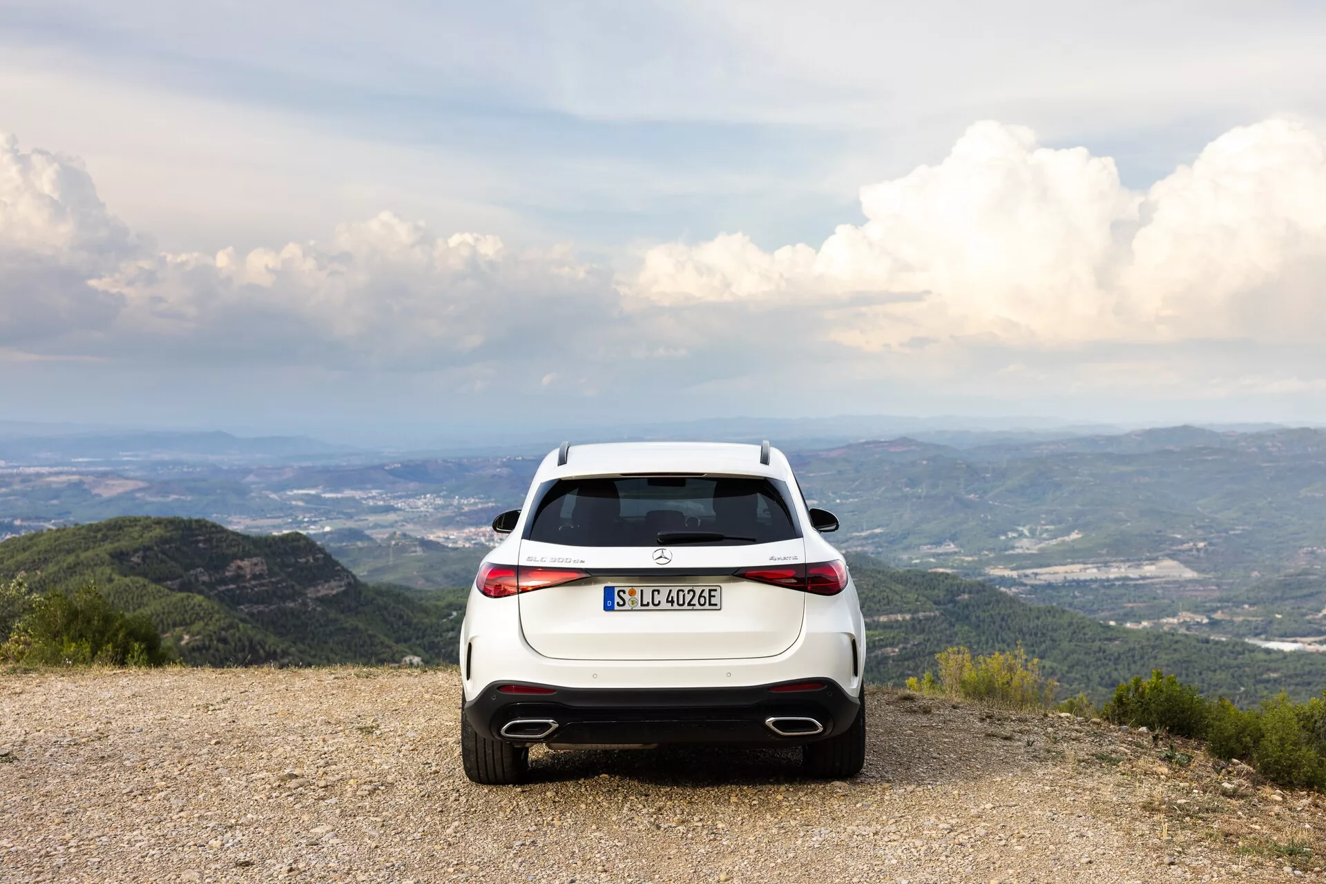 Pogled od zadaj na bel Mercedes-Benz GLC 2022 (EQ) hibridni/električni SUV, parkiran na gramozni površini s slikovito kuliso gora in dreves pod delno oblačnim nebom. Fotografija poudarja vodoravno usmerjene LED zadnje luči, dvojne izpušne cevi, zadnji difuzor in oznako EQ. Kompozicija nakazuje teme pustolovščine in okolju prijaznih potovanj.