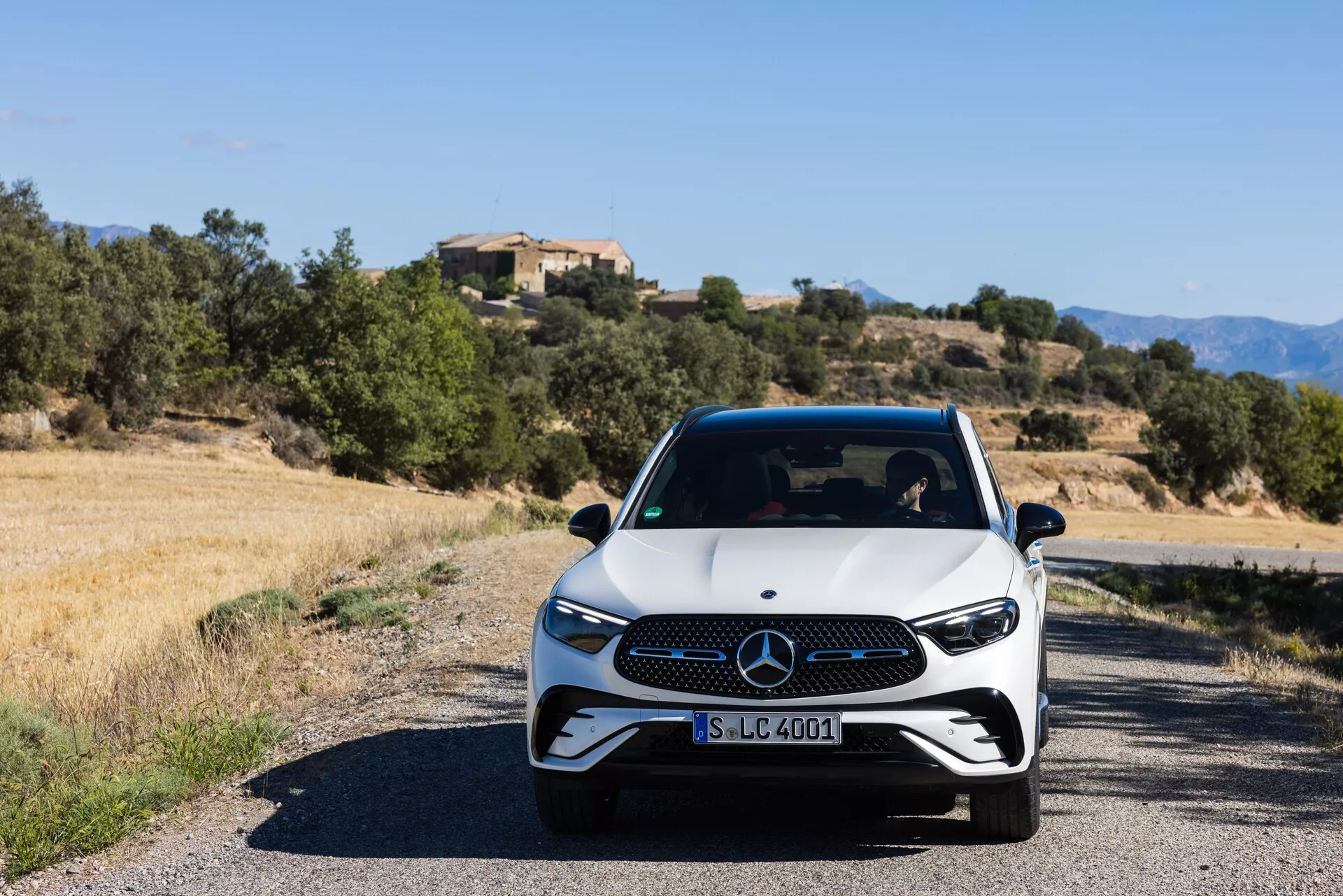 Sprednji kotni pogled na sodoben Mercedes-Benz GLC 2022 (254) na ozki cesti, ki ima belo osnovo s črnimi detajli, LED žaromete in prepoznaven žar. Fotografija, posneta na višini oči podnevi, prikazuje športen in premium videz vozila s sredozemsko pokrajino v ozadju.