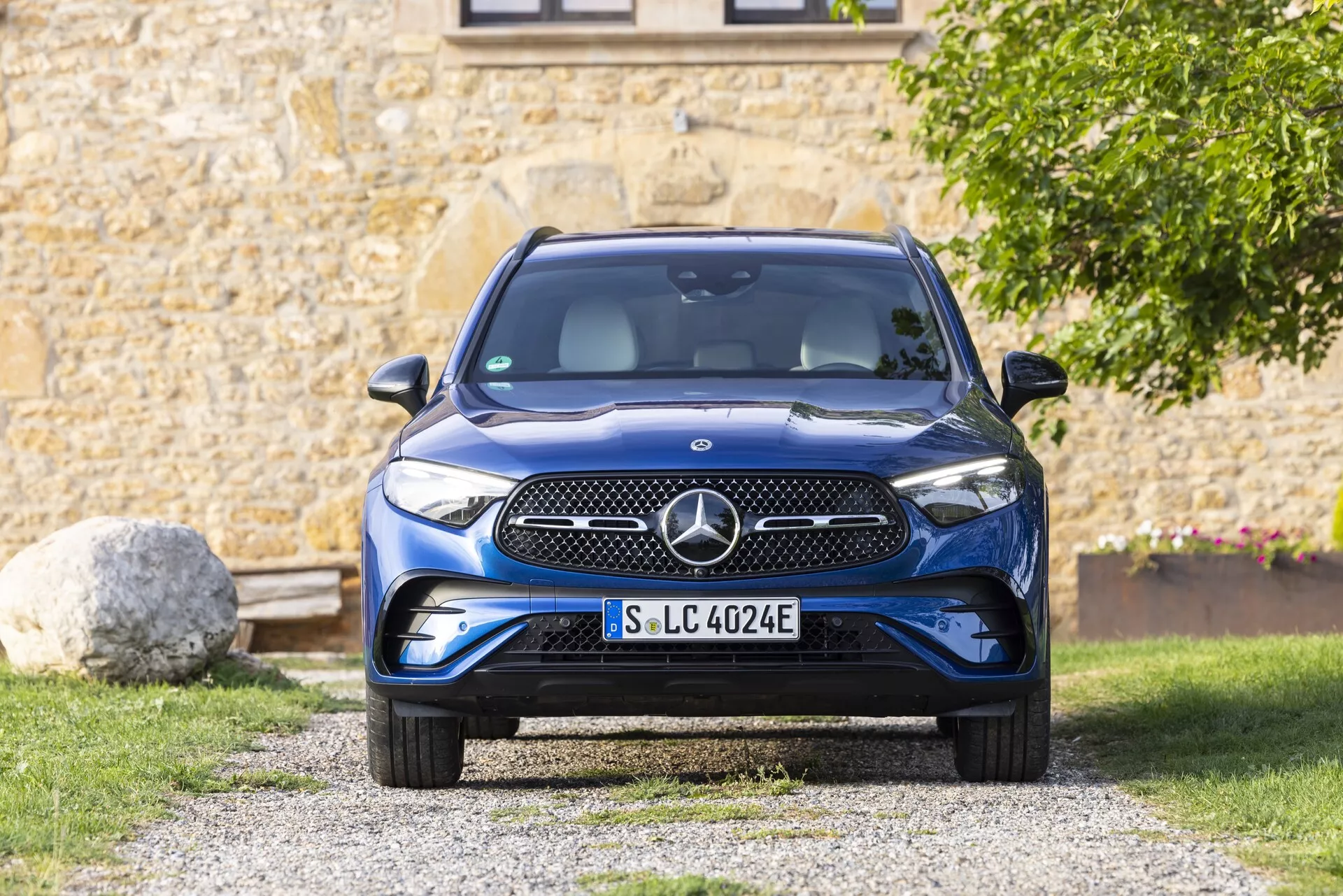 Frontalni pogled na Mercedes GLC 2022 (254), ki prikazuje izrazito prednjo masko, odbijač, pokrov motorja, žaromete in prednje vetrobransko steklo. Avto je postavljen na dovozu v predmestnem okolju z zelenjem in delom stavbe v ozadju. Maska ima gladko površino z emblemom, ki verjetno vsebuje senzorje ali kamero. Elegantne žaromete imajo tehnologijo LED. Spodnji odbijač vključuje dovode zraka in parkirne senzorje. Modri dodatek na registrski tablici nakazuje, da je to električno vozilo (EV) s specifičnimi funkcijami EV. Veliko vetrobransko steklo nudi dobro vidljivost in verjetno vključuje dežne senzorje in svetlobni senzor. Stranska ogledala, čeprav niso popolnoma vidna, so nameščena na vratih in lahko vključujejo spremljanje mrtvih kotov. Kolesa imajo aerodinamično obliko za izboljšanje učinkovitosti dometa. Slika je dobro osvetljena z naravno svetlobo, kar poudarja podrobnosti in barvo avtomobila.