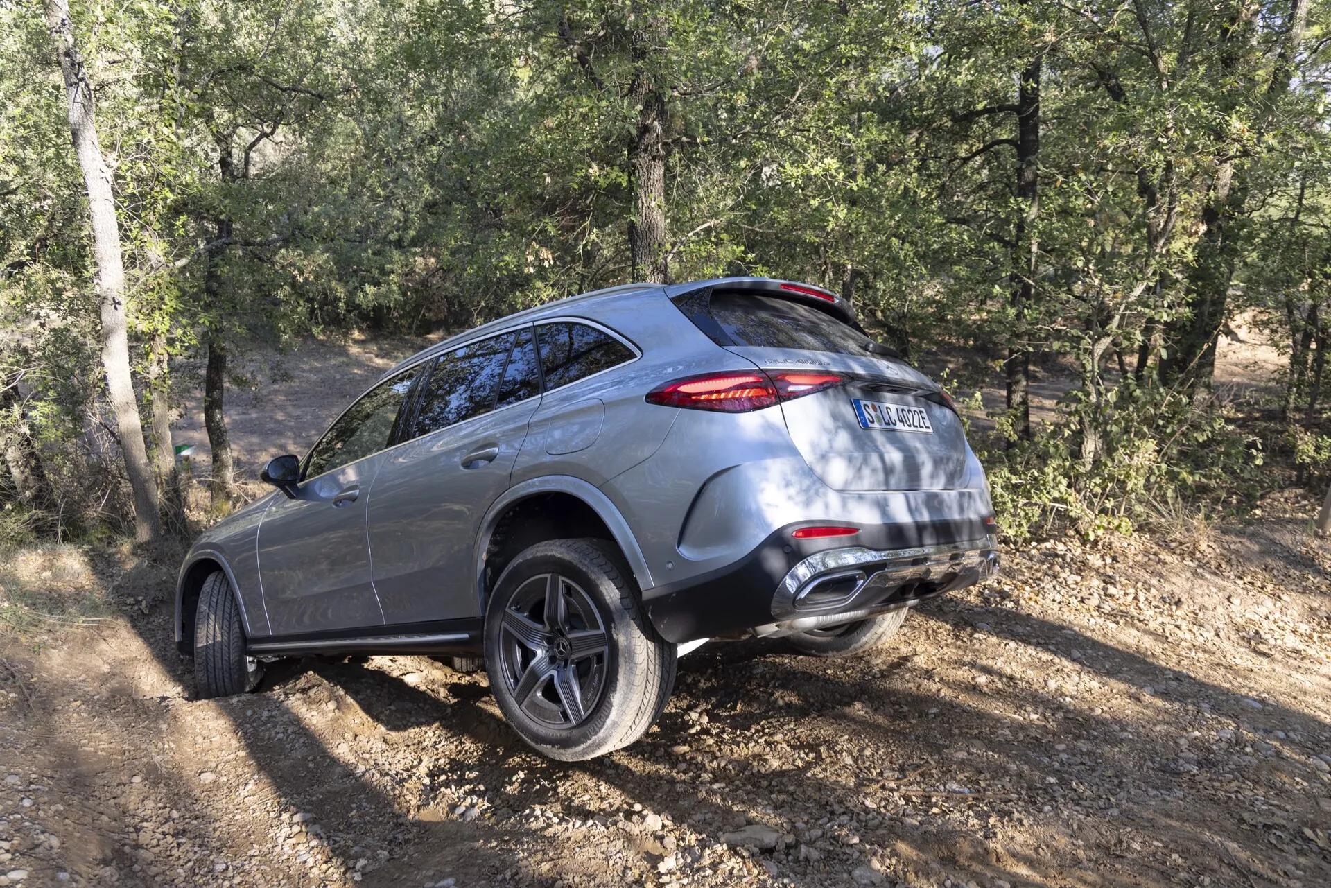 Zadnji trije-četrtinski pogled na SUV Mercedes GLC 2022 na neasfaltiranem terenu, ki prikazuje njegove luksuzne in moderne oblikovalske lastnosti, kot so aluminijasta platišča z več prečkami, LED zadnje luči, dvojni izpušni sistem in terenske zmogljivosti s ozadjem listja in dreves.