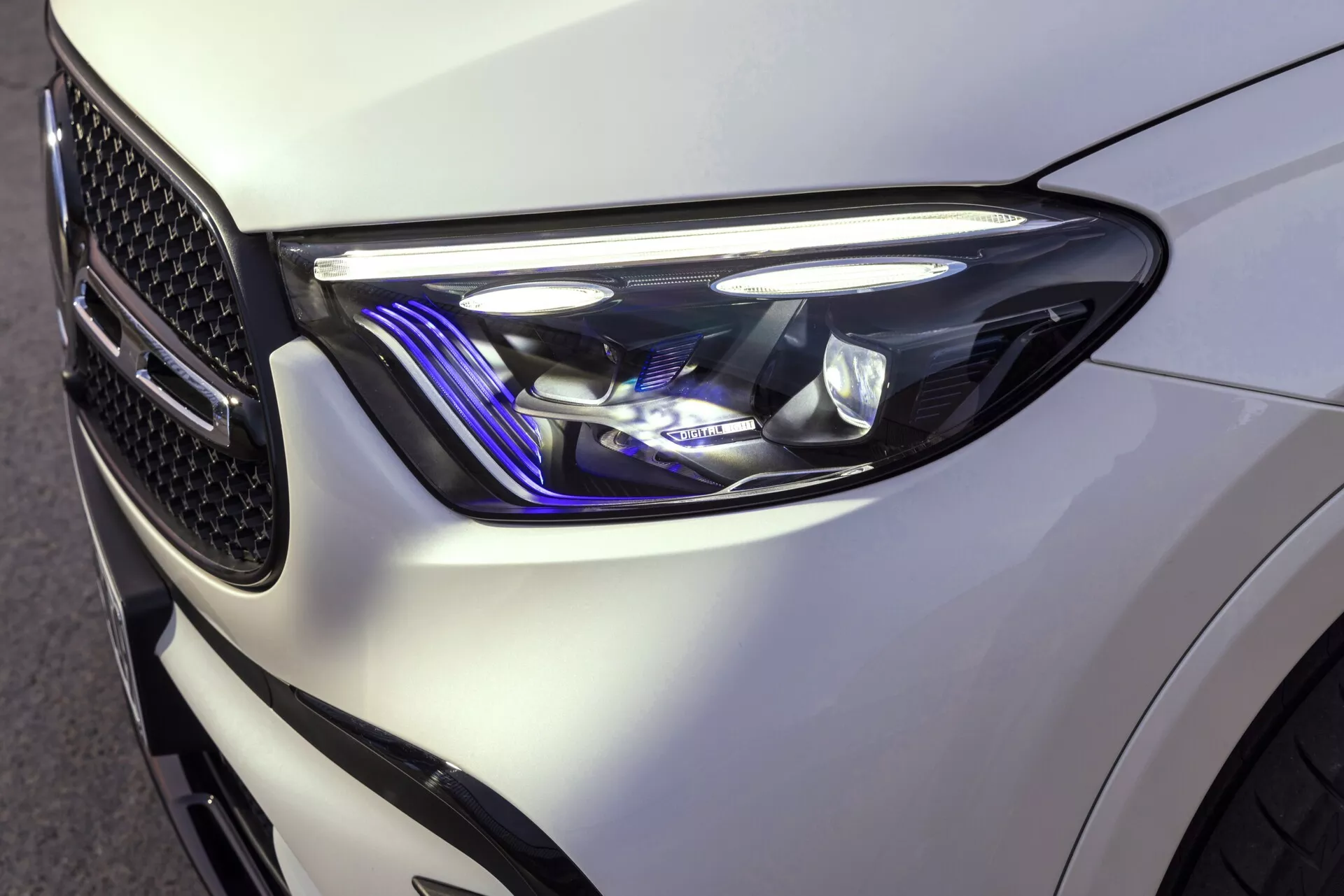 Povečava slike sprednjega levega kvadranta Mercedesa GLC 2022, ki prikazuje eleganten, sodoben dizajn žarometov z ostrimi, kotnimi oblikami in prefinjenimi svetlobnimi elementi, vključno z LED dnevnimi lučmi in modrim poudarkom. Sijajna bela barva poudarja dobro definirane linije vozila, vidna mreža v obliki satja pa odseva njegovo zmogljivost usmerjeno oblikovanje.