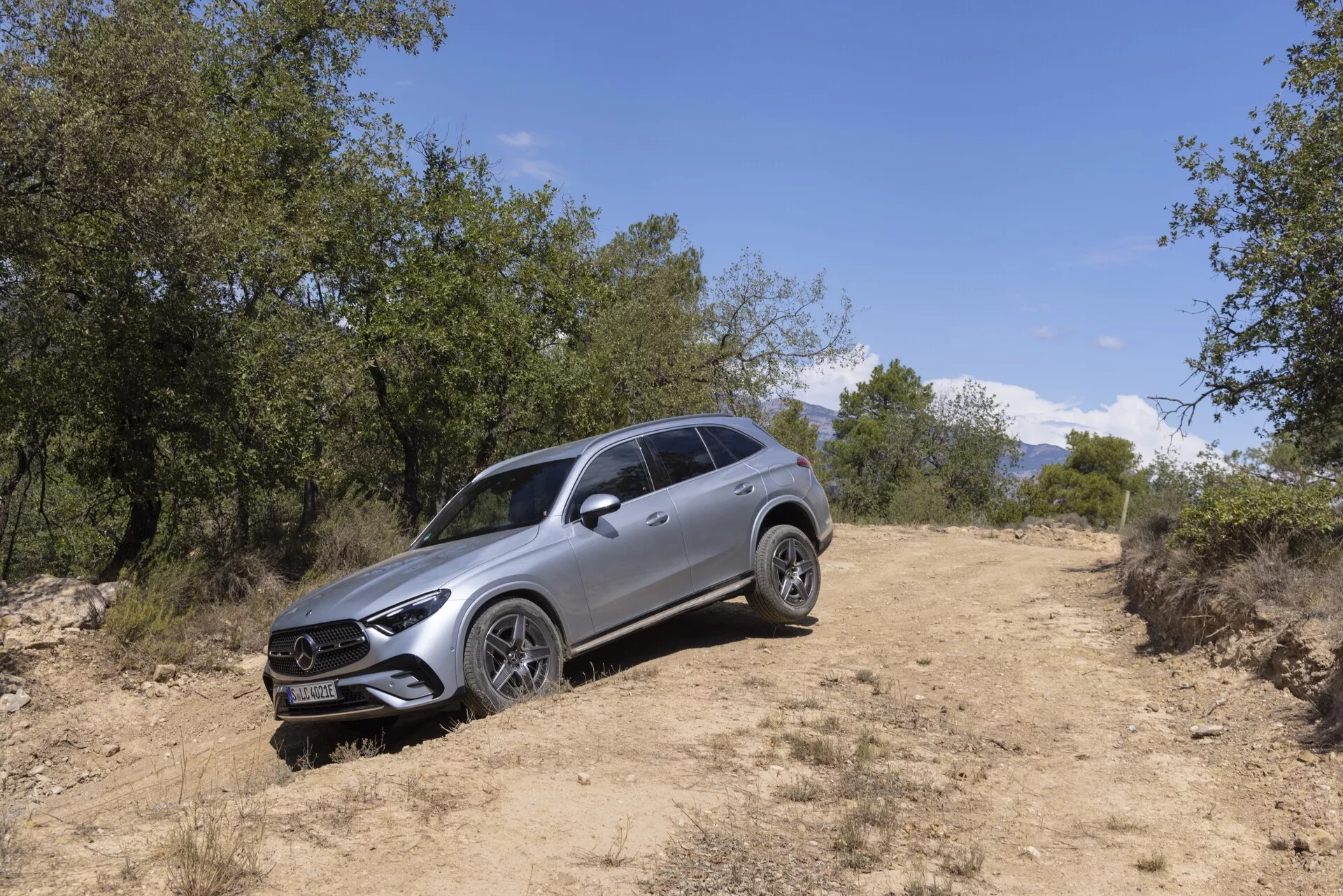Slika SUV-ja Mercedes GLC 2022 v naravnem terenskem okolju, posneta pod kotom treh četrtin z leve prednje strani. Vidni elementi vključujejo izrazito mrežo z osrednjim emblemom, LED žaromete z dnevnimi lučmi, večkrake aluminijaste platišč, strešne tirnice, oblikovano karoserijo, stranska ogledala z integriranimi smerniki ter višjo oddaljenostjo od tal, kar nakazuje na terenske zmogljivosti. Vozilo je postavljeno na makadamski poti, obdano s travo in drevesi, naravna sončna svetloba pa osvetljuje njegov sodoben dizajn in luksuzne lastnosti.