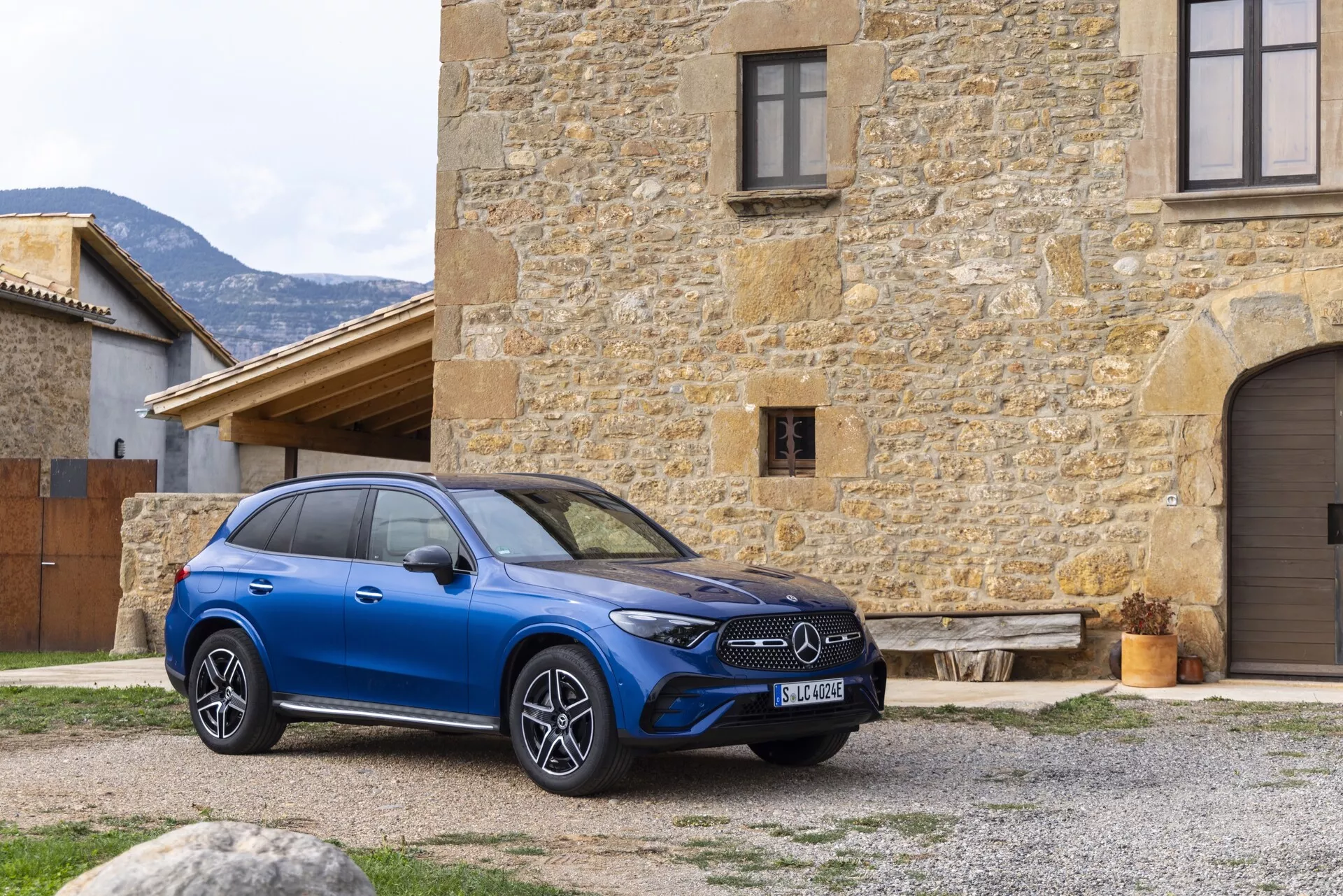 Moderen modri Mercedes GLC 2022 SUV je izrazito prikazan na prostem, postavljen pod rahlim kotom iz središča, kar poudarja sprednjo desno četrtino in del stranskega profila vozila. Avto krasi izrazita sprednja rešetka z emblemi proizvajalca, elegantne vodoravne LED luči in velika večkraka aluminijasta platišča. Je postavljen pred staro kamnito stavbo, kar zagotavlja izrazit kontrast. Potencialne tehnološke lastnosti, kot so adaptivni tempomat, pomoč pri ohranjanju smeri vožnje in panoramska streha, so nakazane v njegovi zasnovi. Sestava slike ponuja jasno, naravno perspektivo luksuznih in visokotehnoloških atributov vozila ter ujame podroben zunanji del v rustikalnem okolju.