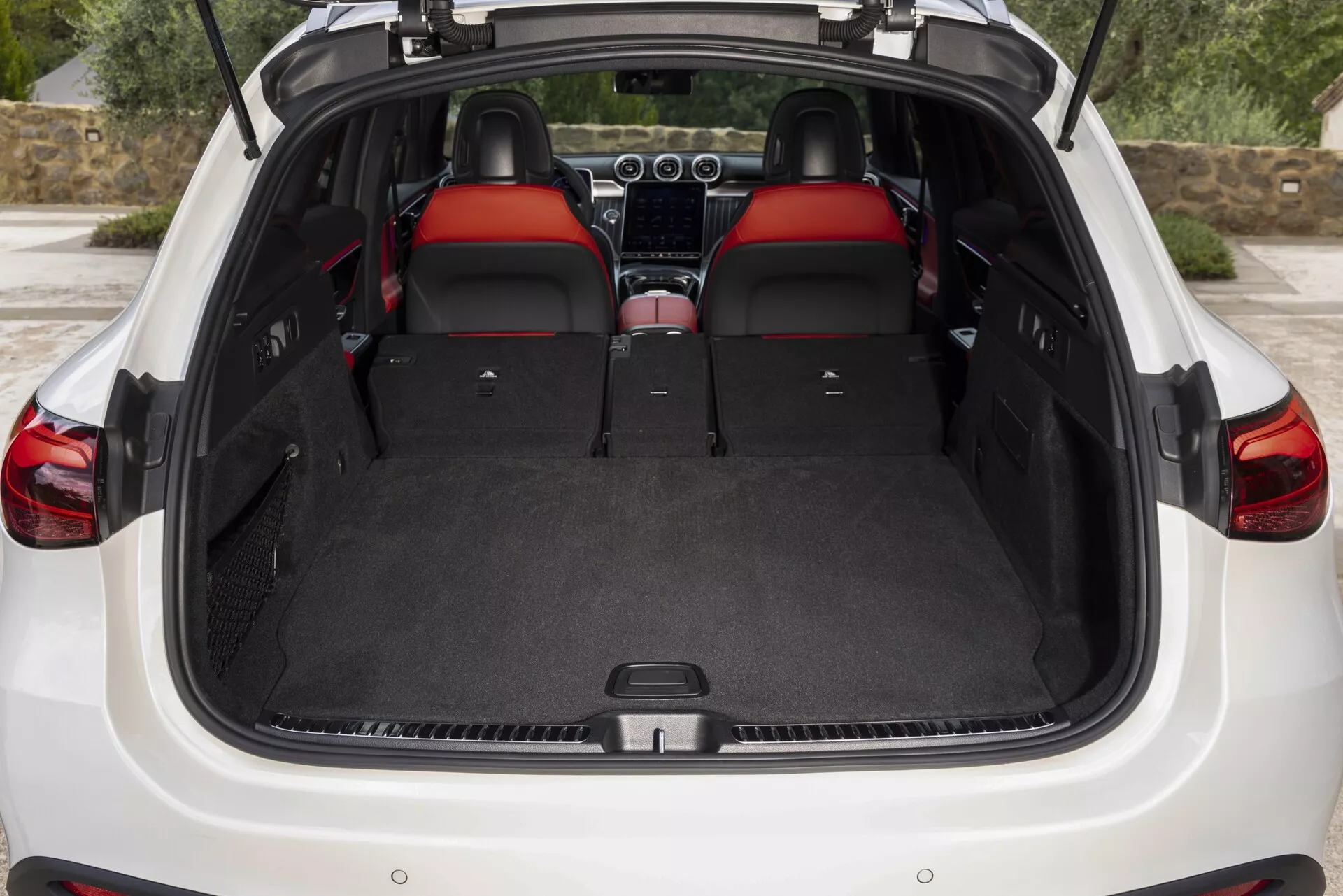 Slika prikazuje zadnji del notranjosti Mercedes GLC 2022 (254) z odprtim prtljažnikom. Pogled ponuja jasen pogled na prostoren, tepihom prekrit tovorni prostor, s ploščatim tovorom tal, kar poudarja prilagodljivost prostora z zadnjimi sedeži, zloženimi v konfiguraciji 60/40. Notranjost ponuja premium rdeče usnjene sedeže, naslonjala za glavo z rdečimi vložki in kontinuiteto dizajna. Delno so vidni armaturna plošča in sredinska konzola, kar daje slutiti dodatne funkcije, kot so infozabavni in klimatski sistemi. Zadnja vrata nakazujejo uporabniku prijazen dizajn, ki lahko vključuje tehnologijo, kot sta brezžično ali električno odpiranje. Pokrova tovora ni prisoten, kar bi lahko pomenilo, da je zložljiv ali odstranjen.