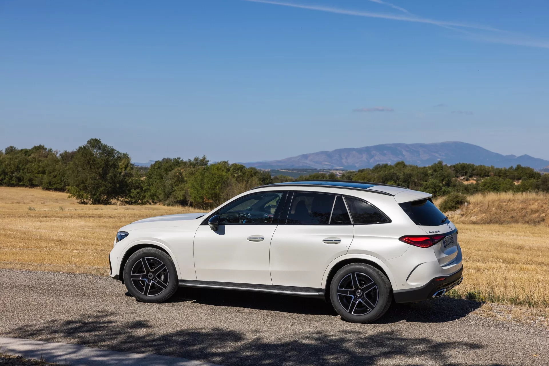 Sodoben luksuzni crossover SUV v podeželskem okolju: tričetrtinski zadnji bočni pogled na svetlo Mercedes GLC 2022 (254) s športnimi temnimi kolesi, toniranimi stekli, poševno strešno linijo in oblikovanim zadnjim odbijačem, parkiran v bližini suhe travnate poljane in dreves v jasnem dnevu.
