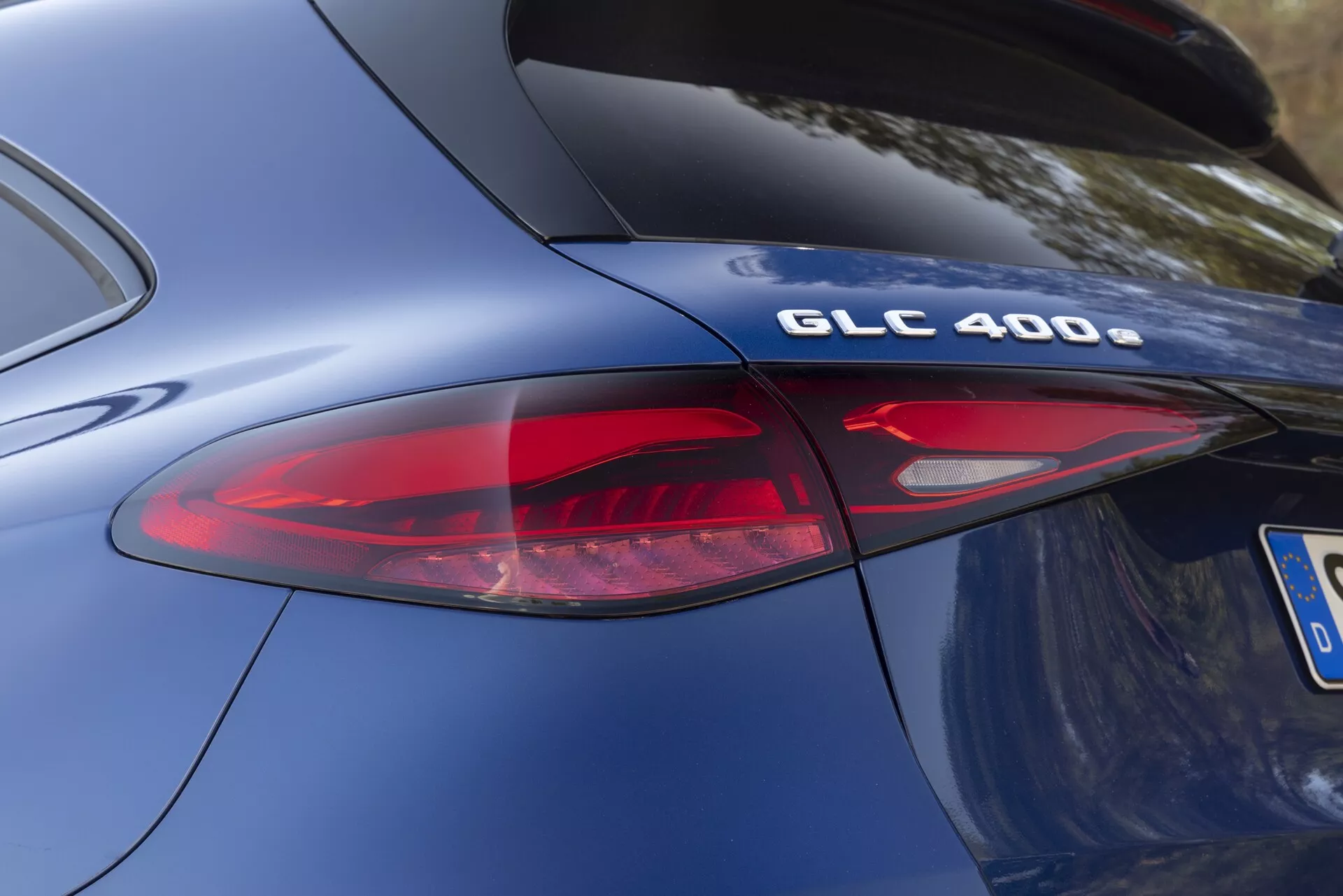 Bližnji, poševen pogled na zadnji del modela Mercedes-Benz GLC 400 2022. Osredotoča se na oznako modela 'GLC 400' in del sodobne LED zadnje luči. Fotografija poudarja krivine karoserije avtomobila, gladko metallicno modro barvo in eleganten dizajn zadnjih elementov, vključno z rdečimi in jasnimi lečami zadnje luči. Kromirana obdelava na znački 'GLC 400' nakazuje specifikacije tehnologije zmogljivosti in pogonskega sklopa.