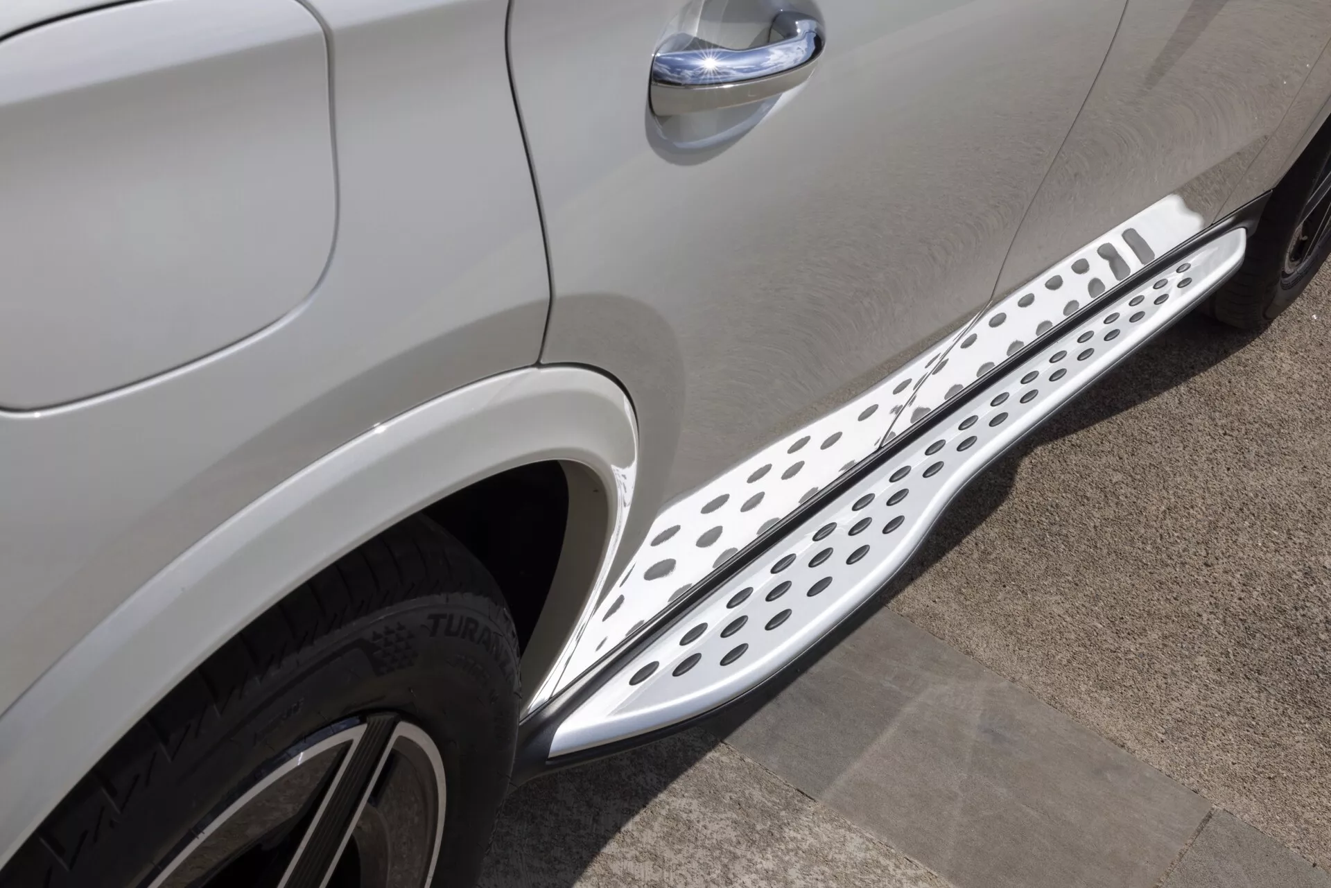Povečan stranski pogled na Mercedes GLC 2022 (254) v kovinsko svetlo sivi ali srebrni barvi, poudarjajoč spodnji del med prednjimi in zadnjimi kolesi. Vidni so del prednjih potniških vrat in del zadnjih vrat, kar nakazuje štirivratni model. Kovinska bočna stopnica s krožnimi izrezi, najverjetneje aluminijasta ali iz nerjavečega jekla, teče po dolžini med koloteki. Fotografija prikazuje tudi zgornji del prednje pnevmatike na potniški strani s tekstom Bridgestone TURANZA. Dodatne podrobnosti vključujejo kromirano obrobo na kljuki vrat in linijo oblikovanja karoserije, kar kaže na luksuzno opremo in SUV oblikovanje.