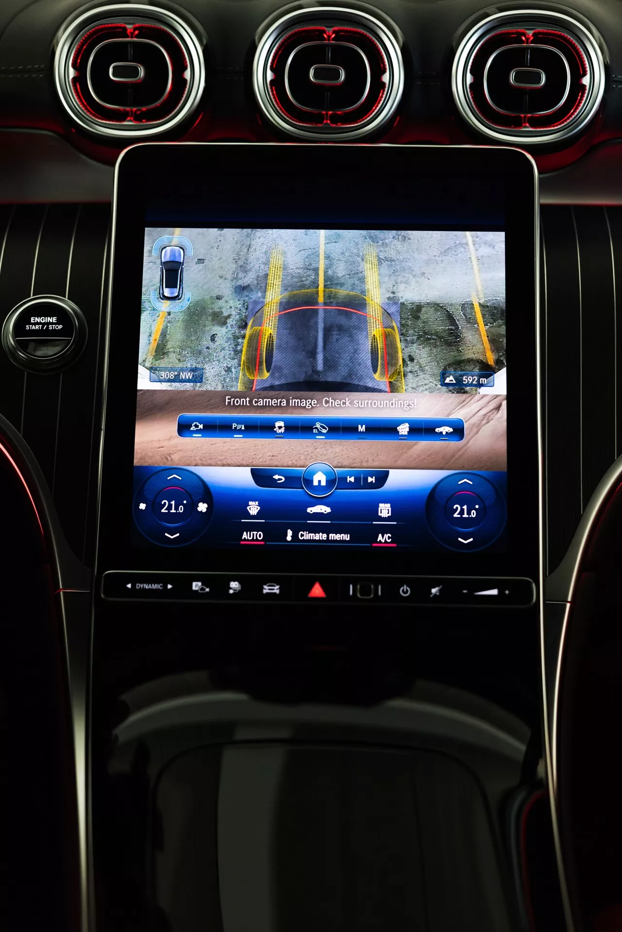 Notranjost Mercedes GLC 2022 s centralno konzolo s zaslonom na dotik infotainmenta, ki prikazuje sliko prednje kamere z rumenimi in modrimi linijami za pomoč pri parkiranju. Pogled s sedeža voznika vključuje prezračevalne reže z rdečimi detajli nad zaslonom ter gumbe za upravljanje klime pod njim.