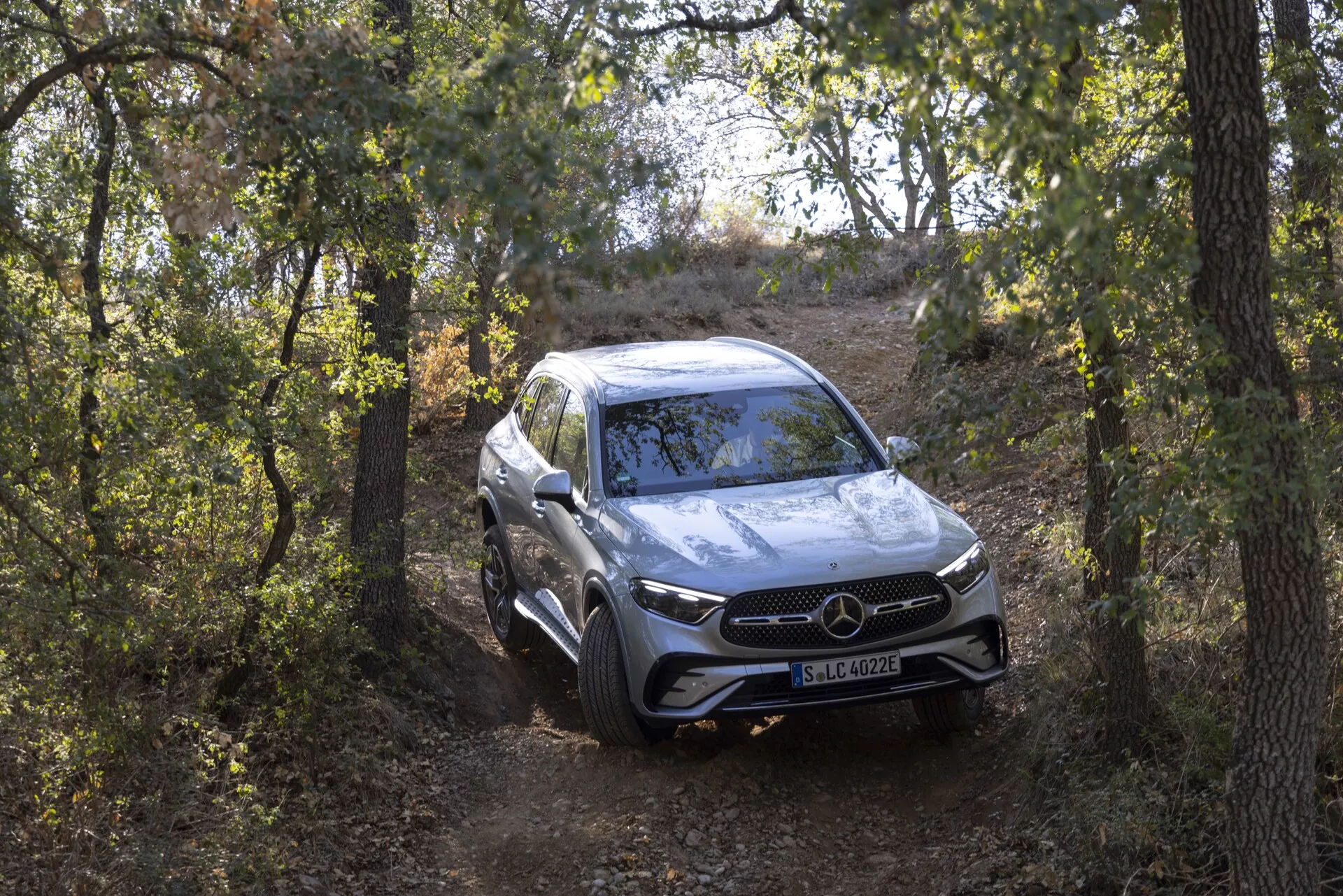 Celoten pogled na SUV Mercedes GLC 254 iz leta 2022 iz sprednje tričetrtinske perspektive, ki se približuje kameri na naravni poti, obdani z drevesi. Rahlo visok kot poudarja njegove terenske zmogljivosti, višino vožnje in nastavitev vzmetenja. Luksuzno vozilo ima izrazito sprednjo mrežo z velikim emblemom, LED žaromete, parkirne senzorje in aerodinamične konturirane črte na pokrovu motorja. Srebrna barva odraža okolico, medtem ko aluminijasta platišča in vsestranski tekalni profil dodajajo športnemu in elegantnemu videzu.