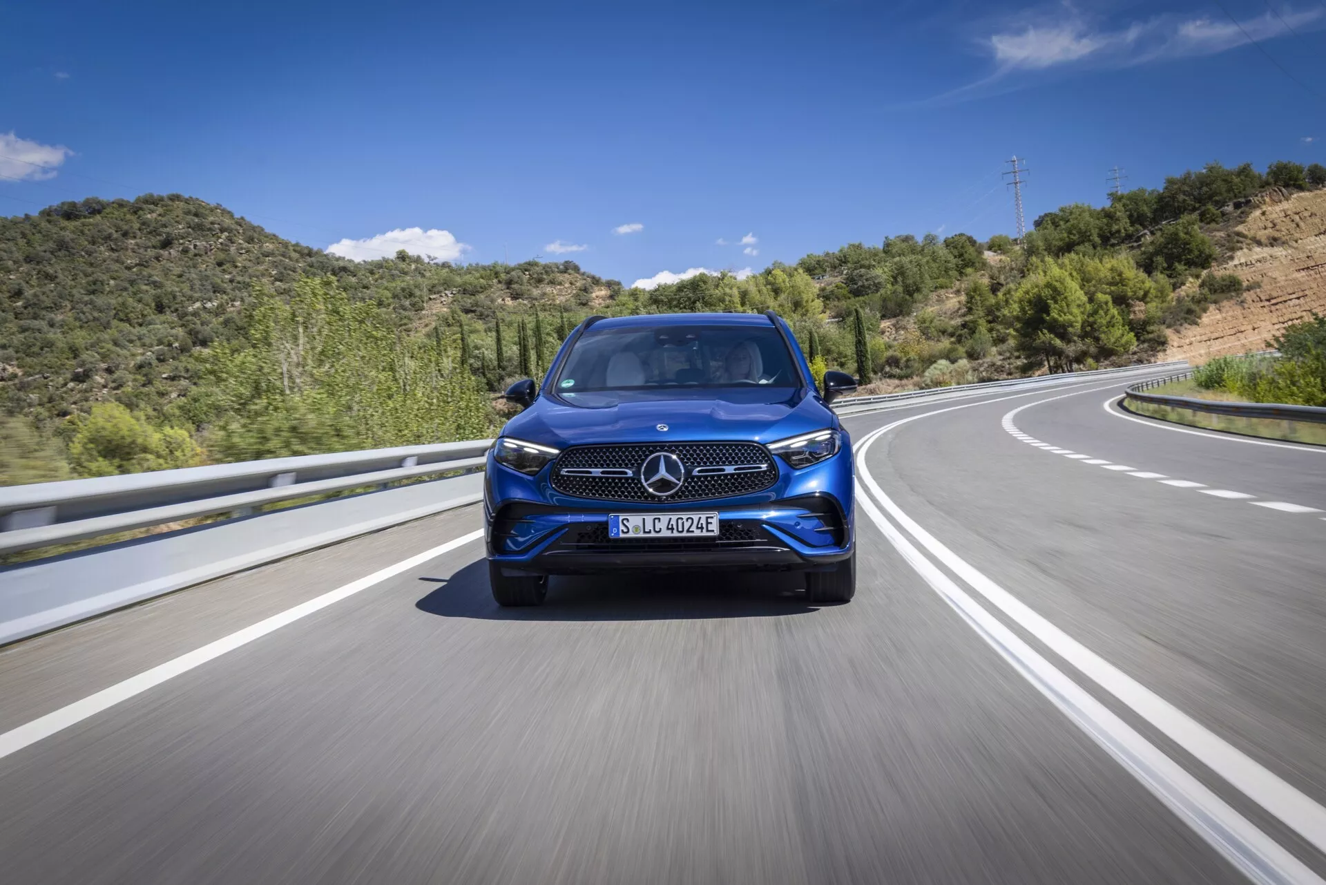 Nizko kotni, čelni pogled na moderen Mercedes GLC 2022, ki vozi po avtocesti ali podeželski cesti. Ta slika poudarja prepoznavno mrežo vozila, obliko žarometov, stil sprednjega odbijača, velika lita platišča in potencialne napredne sisteme za pomoč voznikom (ADAS). Osvetlitev je svetla in naravna s ozadjem modrega neba in zelenih gričev, kar namiguje na gibanje po asfaltu z ločilnimi oznakami pasov.