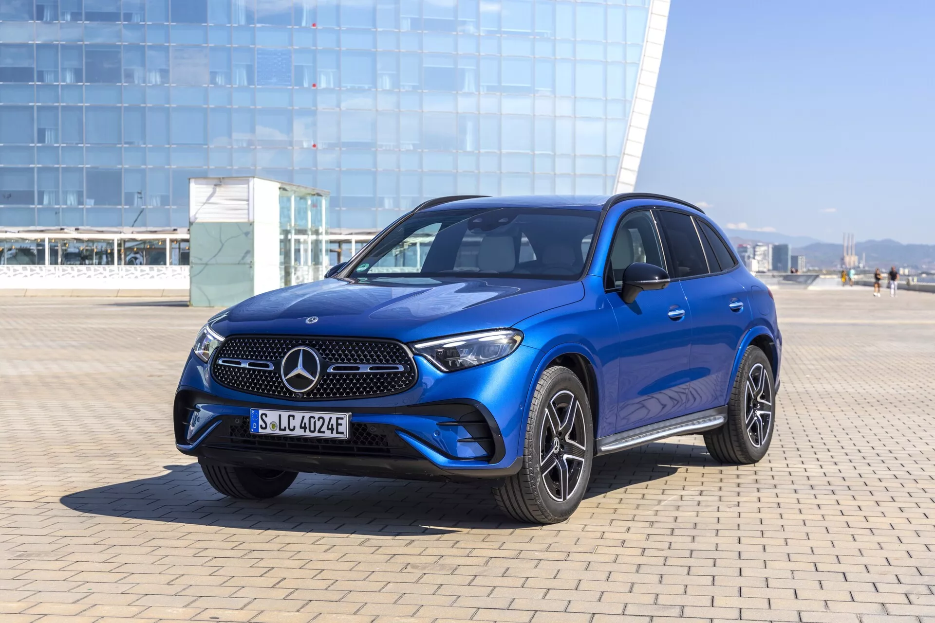 Sprednji trikotni pogled na moder Mercedes-Benz GLC SUV 2022, ki prikazuje značilno Mercedesovo masko z logotipom, elegantne vodoravne letvice, integrirani odbijač z meglenkami in zračniki, ostre LED žaromete, oblikovane črte pokrova motorja, športna večkraka platišča iz lahke litine, kljuke in ogledala v barvi karoserije, zatemnjena okna, strešno okno in možne napredne sisteme za pomoč vozniku. Fotografirano v svetlem zunanjem okolju z naravno sončno svetlobo in čisto modrim nebom.