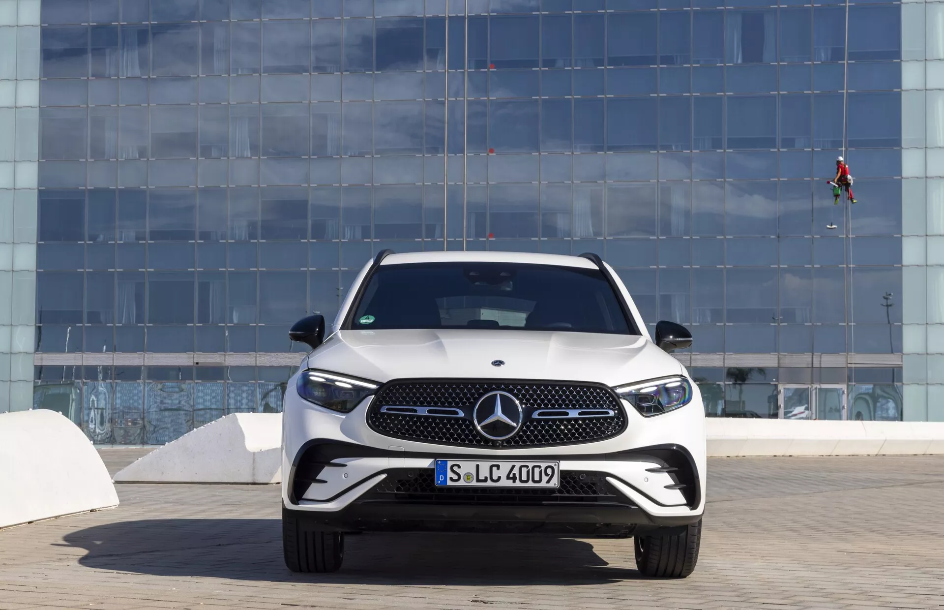 Sprednji pogled na Mercedes-Benz GLC 2022 (254), ki prikazuje podroben dizajn sprednjega dela, rešetko z izrazitim emblemom Mercedes, elegantne LED žaromete, športni odbijač z velikimi odprtinami za zrak in aerodinamičnimi dodatki, v mestnem okolju na svetel dan.