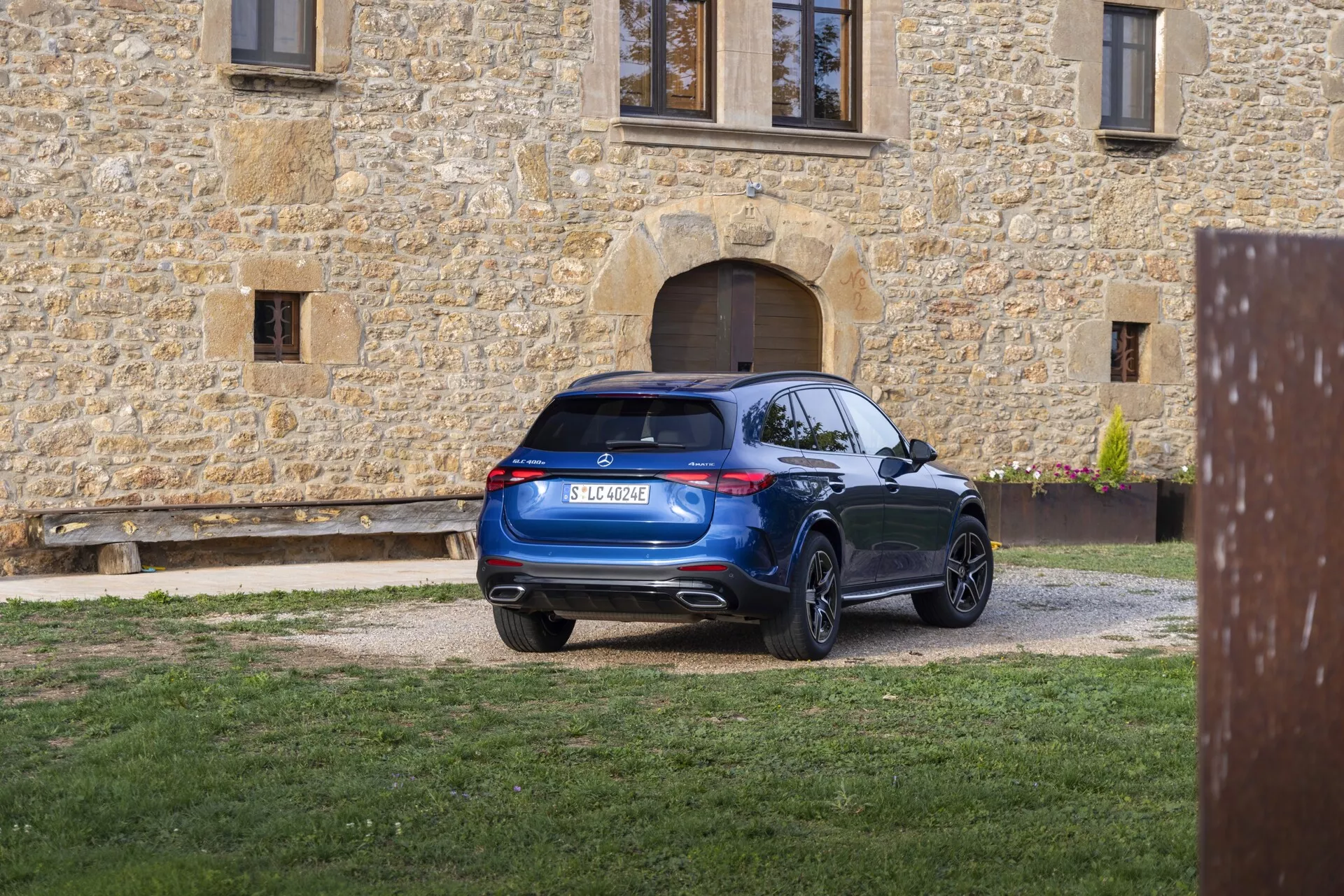 Moderen SUV parkiran na gramozni dovozni poti, viden iz treh četrtin zadnje perspektive. Zadnje luči so opremljene s sodobno LED tehnologijo, ki je gladko integrirana v dizajn. Zadnje steklo, rahlo nagnjeno naprej, prispeva k aerodinamičnemu profilu, ki ga dopolnjuje strešni spojler. Zadnji odbijač ima vgrajene senzorje ali odsevnike, kar nakazuje na parkirni sistem, dvojni izpušni cevki pa sta vgrajeni v spodnji del odbijača. SUV se ponaša z velikimi, večkrakimi aluminijastimi platišči in površino v bogatem modrem odtenku, ki odraža dobro vzdrževan videz proti ozadju rustikalne kamnite stavbe.