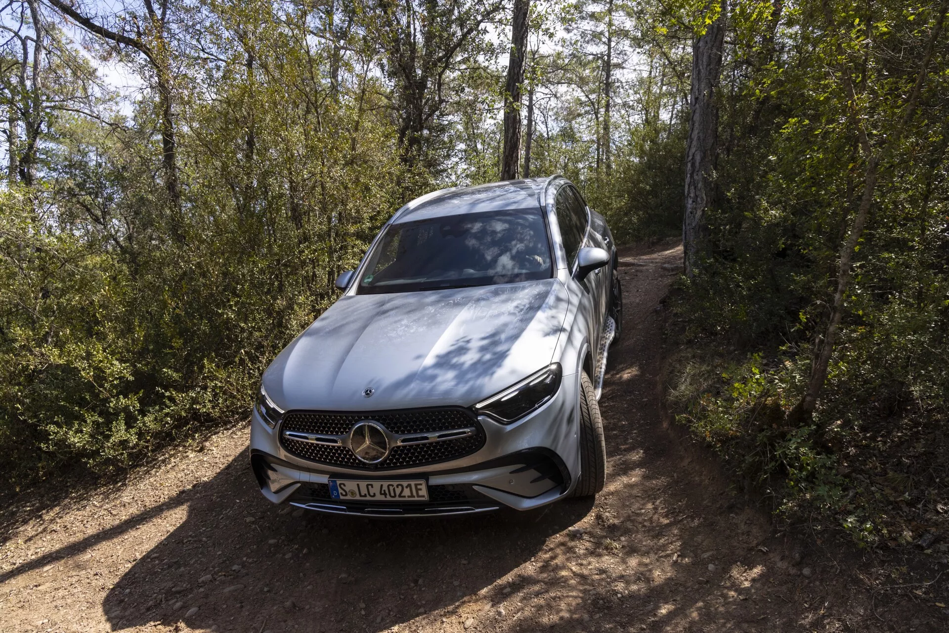 Sodobni luksuzni avto, Mercedes-Benz GLC 2022, visok sprednji kot perspektive, ki prikazuje sprednjo masko, odbijač, pokrov motorja, levo stran vključno z voznikovo vrati in bočnim ogledalom, eleganten sodoben dizajn, poudarjen trikraki znak Mercedes-Benz, podrobne LED žaromete, večkraka aluminijasta platišča, možen znak EQ, ki označuje električni ali hibridni model, verjetno funkcije ADAS, nameščen na makadamski cesti obdano z vegetacijo, srebrna ali svetlo siva kovinska barva, ki poudarja vrhunsko kakovost.