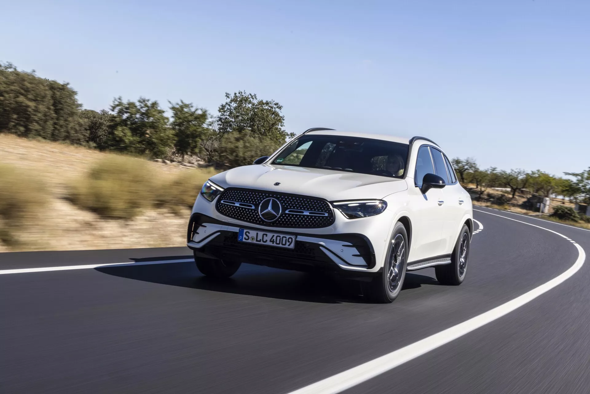 Luksuzni beli Mercedes GLC 2022 crossover SUV vozi po gladki asfaltirani cesti, naravna pokrajina v ozadju pod jasnim dnevnim nebom. Dinamičen posnetek z nizkega kota, ki zajame pogled na sprednjo tretjino z LED žarometi, izrazito sprednjo mrežo z emblemom, aerodinamično zasnovo in elegantnimi lahkimi kolesi, poudarjanje občutka hitrosti in gibanja s rahlim zameglitvijo ozadja.