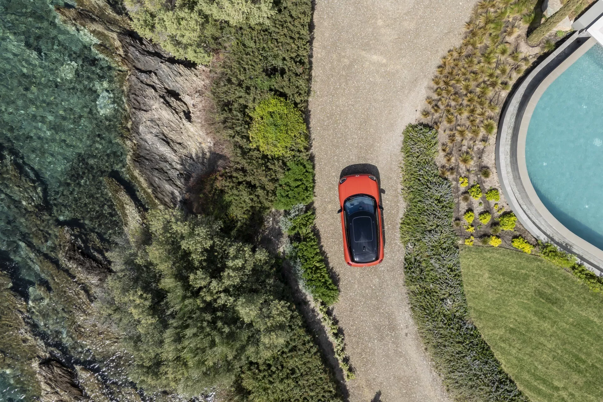 Pogled iz zraka na Mini Aceman 2024 (J05), ki prikazuje njegovo streho, pokrov motorja, prtljažnik in del stranskega profila, parkiran ob pečini in telesu vode, obdan z bujno vegetacijo, poudarjajoč lastnost sončne strehe