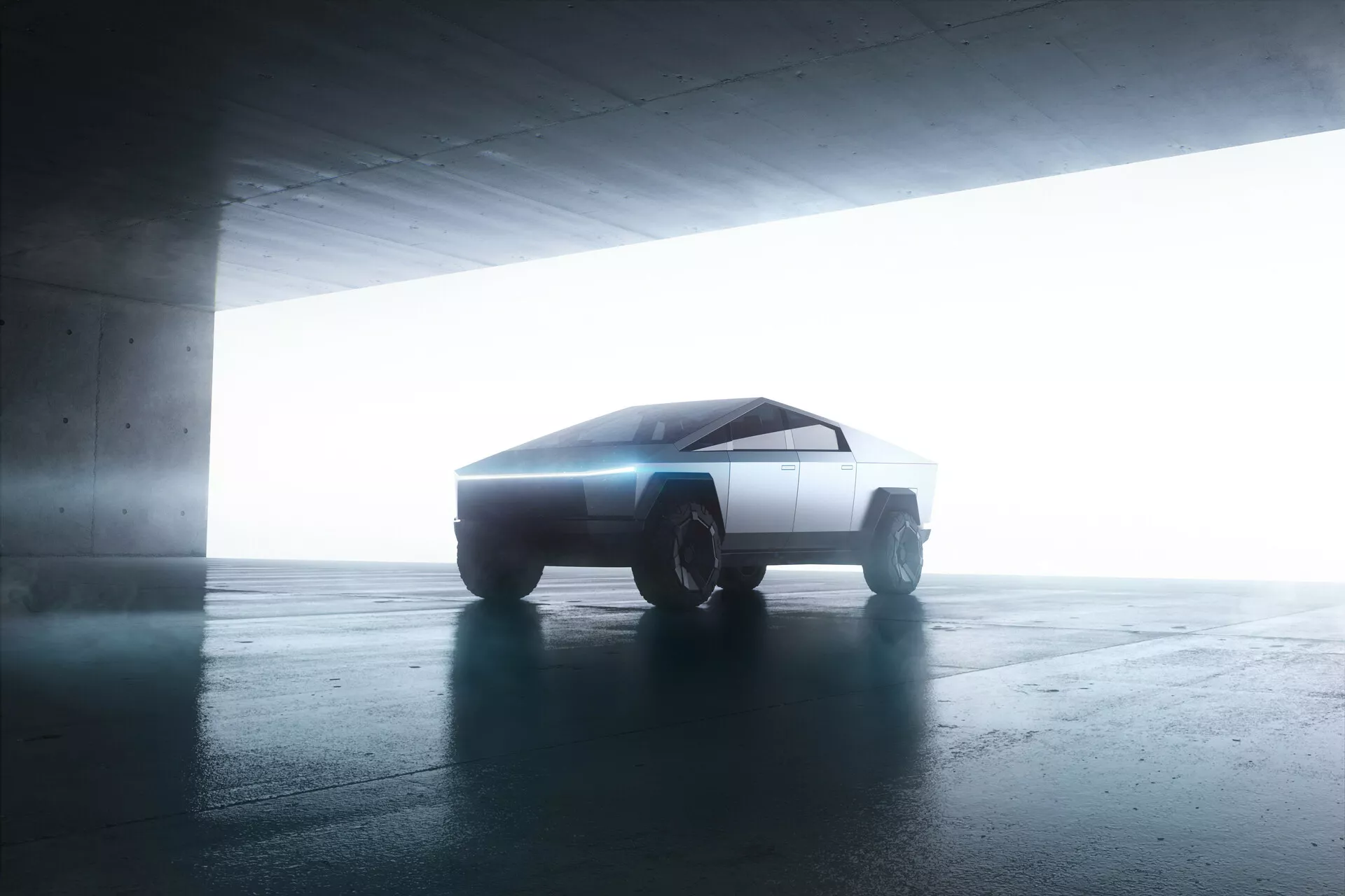Stranski pogled na Tesla Cybertruck 2021 v parkirni garaži. Vozilo s futurističnim, kotnim dizajnom je rahlo nagnjeno proti fotoaparatu, kar razkriva njegove minimalistične, geometrične linije. Osvetlitev od zadaj poudarja njegovo silhueto, ustvarja halo učinek. Ostro in plosko oblikovane površine avtomobila ter robustna terenska kolesa nakazujejo napredno, verjetno električno pogonsko tehnologijo.