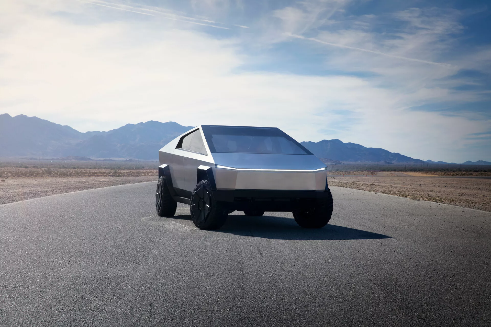 Tesla Cybertruck 2021 (1. generacija), fotografiran iz nizkega sprednjega tričetrtinskega kota, ki prikazuje svojo futuristično, kotno zasnovo. Ta pogled zajame levo sprednjo stran vozila, poudarjajoč njegovo klinasto obliko z ostrimi linijami in ravnimi površinami. Vozilo je opremljeno z velikimi, robustnimi pnevmatikami, primernimi za vožnjo izven ceste, in ima eno samo, neprekinjeno svetlobno črto čez celotno širino sprednje strani, kar kaže na sodobno LED ali lasersko razsvetljavo. Strmo nagnjeno, prostorno vetrobransko steklo in ostro zožena stranska okna prispevajo k njegovi geometrični estetiki. Vozilo je izdelano iz brušenega kovine ali kompozitnega materiala, kar nakazuje vzdržljivost in robustnost. Ozadje vključuje obsežno jasno nebo in gorski teren, kar kaže na suho, puščavsko okolje.