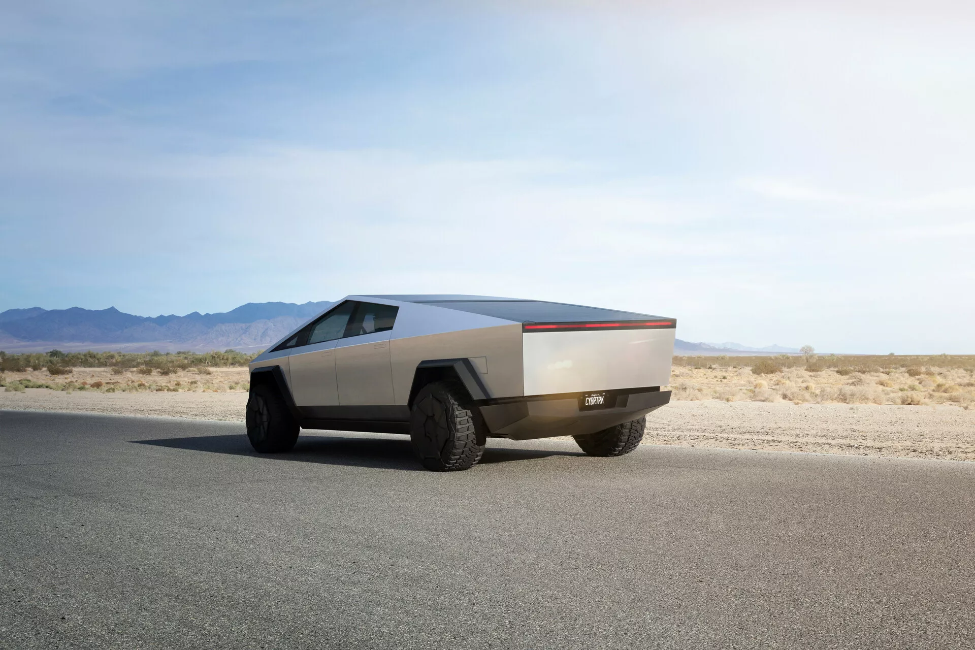 Futuristični Tesla Cybertruck 2021 na ravni asfaltni cesti, posnet iz zadnjega tri četrtinskega kota, poudarja svoj značilen kotni dizajn, kovinsko obdelavo, velike terenske pnevmatike in pomanjkanje tradicionalnih ogledal in kljuk na vratih, kar kaže na napredne avtomatizirane mehanizme.