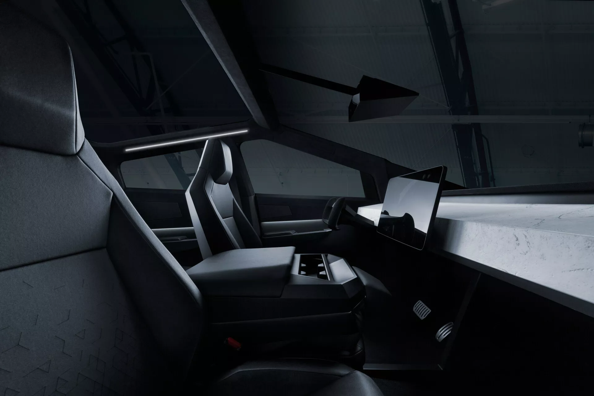 Notranji pogled Tesla Cybertruck 2021 (1. generacija) iz zadnjega sedeža, ki prikazuje desno stran kabine z velikim horizontalno usmerjenim zaslonom na dotik, ki prevladuje na sredini armaturne plošče. Del volana je viden, vendar večinoma prikrit. Sprednji sedeži za sopotnika in voznika imajo integrirane vzglavnike in teksturirano oblazinjenje, verjetno iz blaga ali sintetičnega usnja. Na talnem območju je vidna preproga za avto in niso vidni nobeni ročni kontrolni elementi, kar nakazuje minimalističen, digitalen pristop. Temna barvna paleta in subtilna osvetlitev dajejo moderni, luksuzni videz.